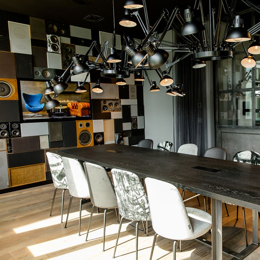 Motel One Besprechungsraum mit langem dunklem Tisch, verschiedenen gepolsterten Stühlen und auffälliger, flexibler Deckenbeleuchtung. Akustisch gestaltete Wand mit Lautsprechern und abstrakten Bildern. Viel Tageslicht durch Fenster reflektiert auf hellem Holzboden.