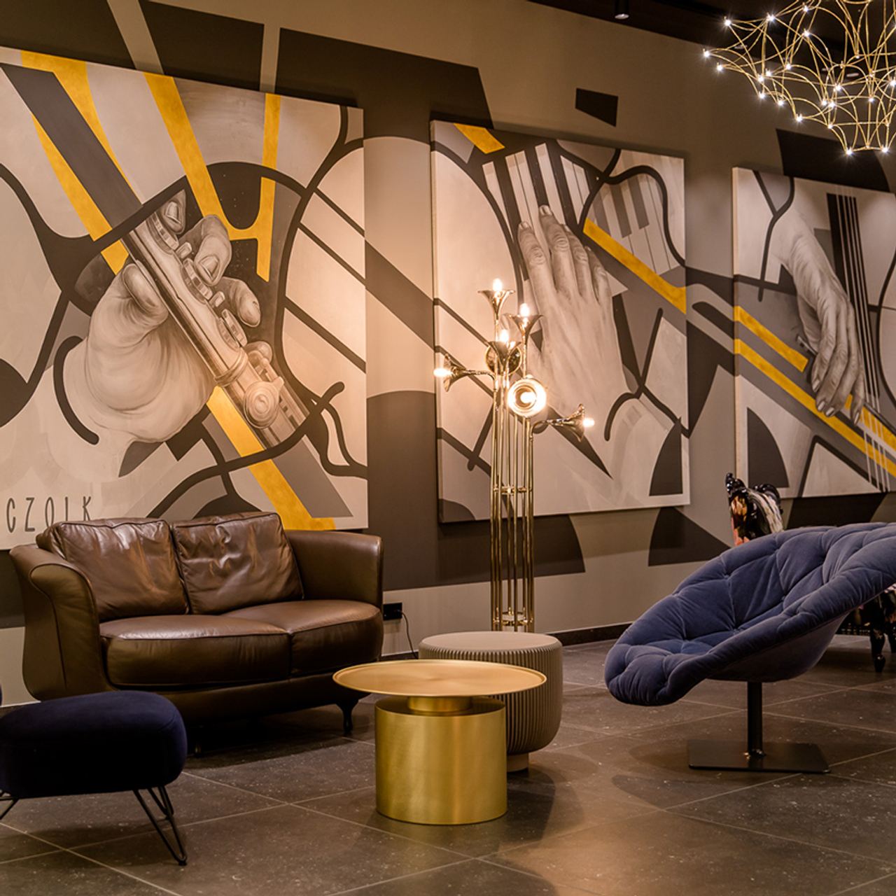 Motel One Lounge-Bereich mit bequemen Sesseln und Couch, stilvollem Beleuchtungskörper und großflächiger Wandmalerei mit detailreichen Händen, die Musikinstrumente spielen.