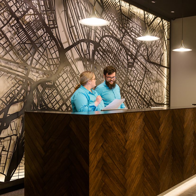 Zwei Motel One Mitarbeiter in blauen Hemden stehen an der stilvollen Rezeption mit dunklem Holz und beleuchtetem Stadtplan an der Wand, im Gespräch und beim Durchsehen von Unterlagen.