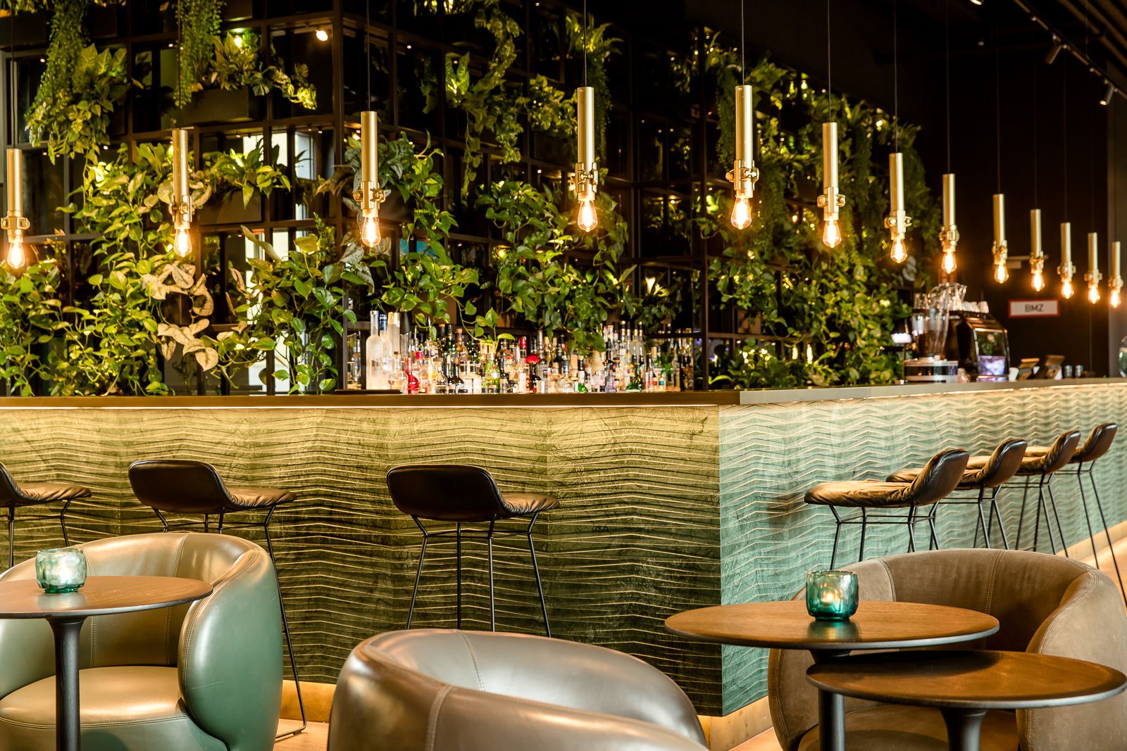Motel One Hotelbar mit stilvoller grüner Wandbegrünung, warmem Licht von hängenden Glühbirnen und Sitzgelegenheiten aus Leder.