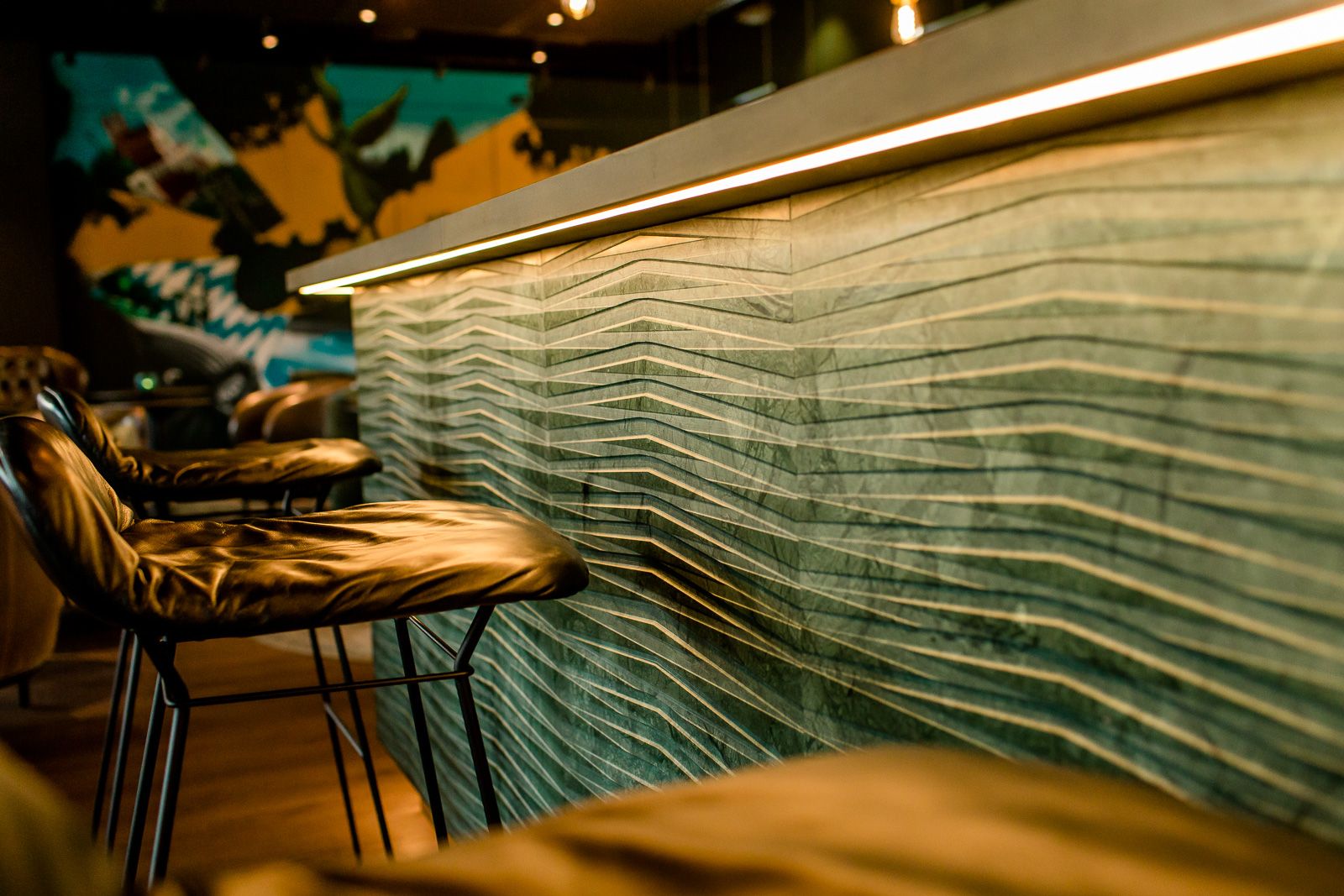 Motel One Hotelbar mit stilvoll gepolsterten Barhockern und moderner, wellenförmiger Wandverkleidung. Sanfte Beleuchtung betont das Design und lädt zu einem gemütlichen Aufenthalt ein.