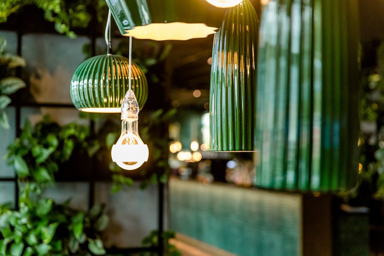 Mehrere grüne, dekorative Pendelleuchten in einem stilvollen, modernen Innenraum, der Wärme und gemütliche Atmosphäre vermittelt. Im Hintergrund sind Pflanzen und sanftes Licht zu erkennen.