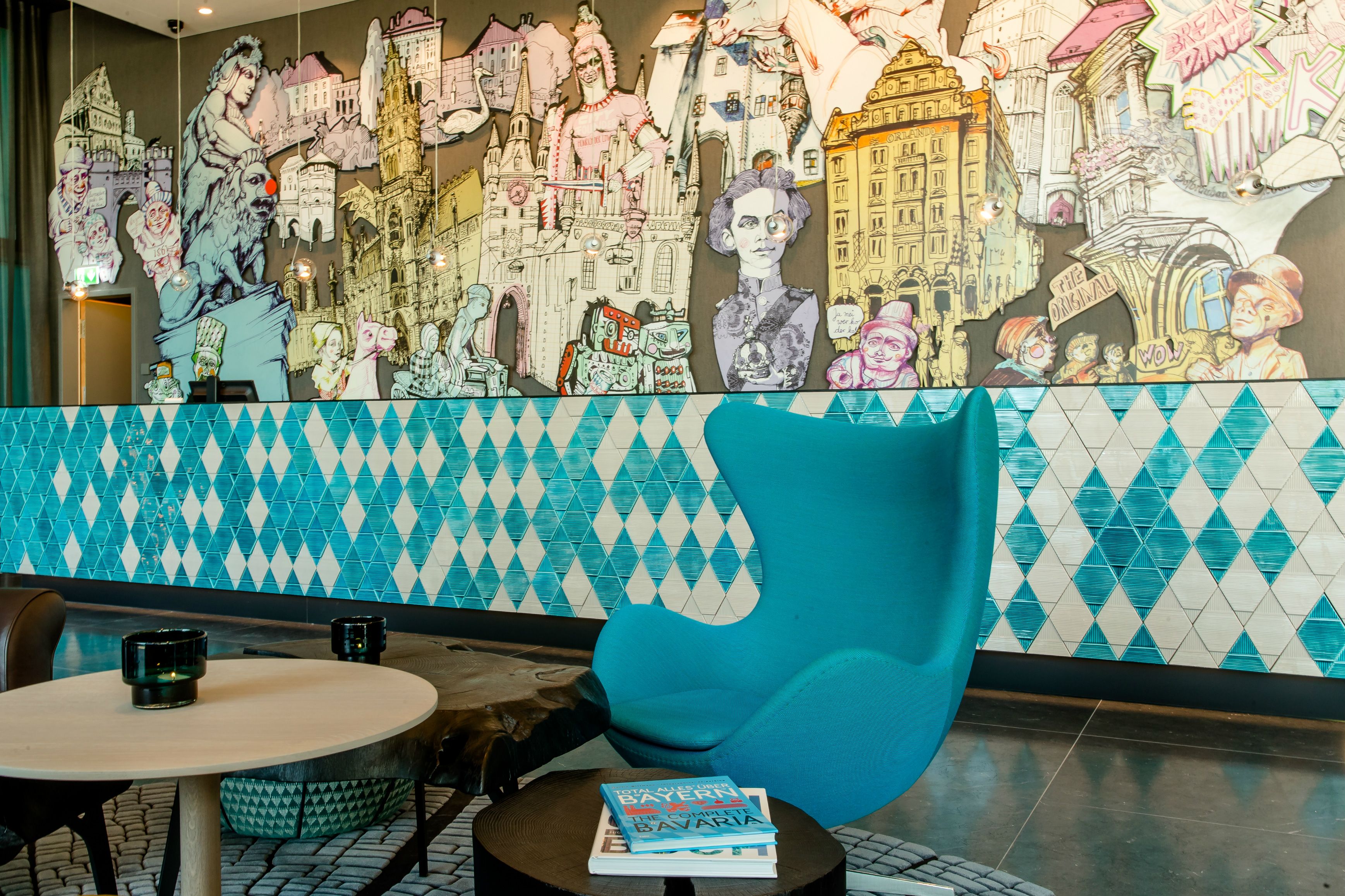 Motel One Lounge-Bereich mit auffälligem Wandgemälde urbaner Motive. Bequemer, türkisfarbener Sessel lädt zum Entspannen ein. Hellblau-weiße, diamantförmige Fliesen


