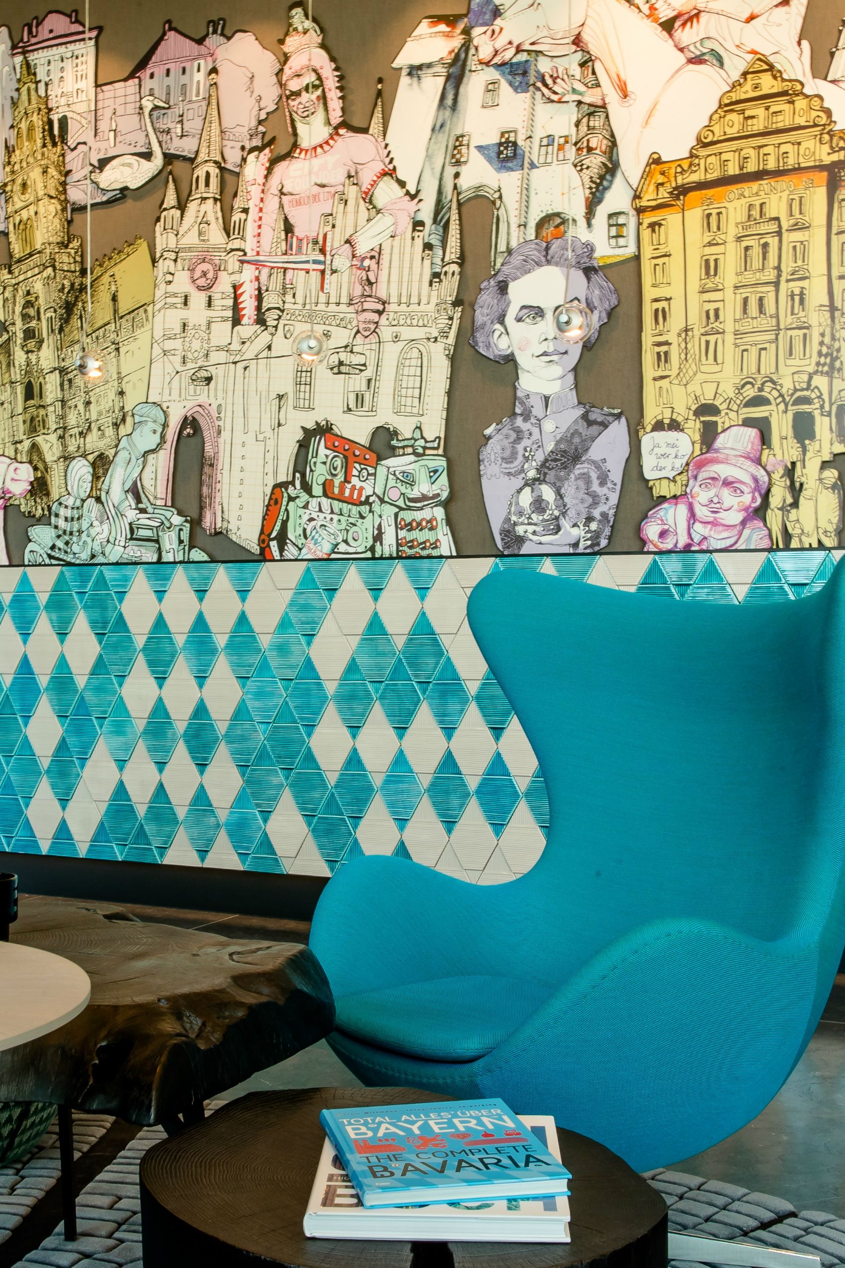 Motel One Lounge-Bereich mit auffälligem Wandgemälde urbaner Motive. Bequemer, türkisfarbener Sessel lädt zum Entspannen ein. Hellblau-weiße, diamantförmige Fliesen