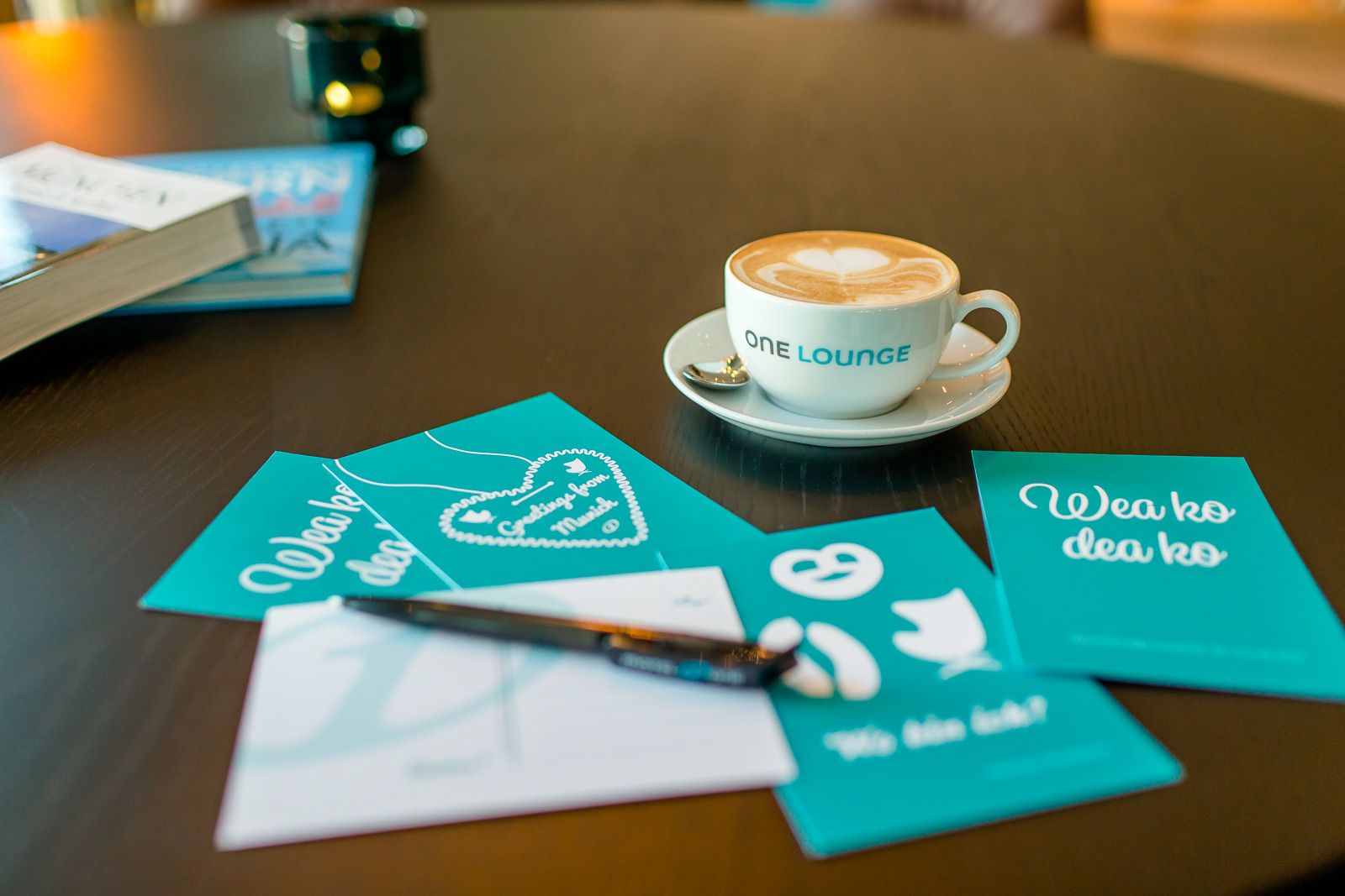 Motel One Tasse mit Kaffee und Milchschaumherz auf Untertasse, umgeben von Grußkarten und einem Stift auf einem Tisch.