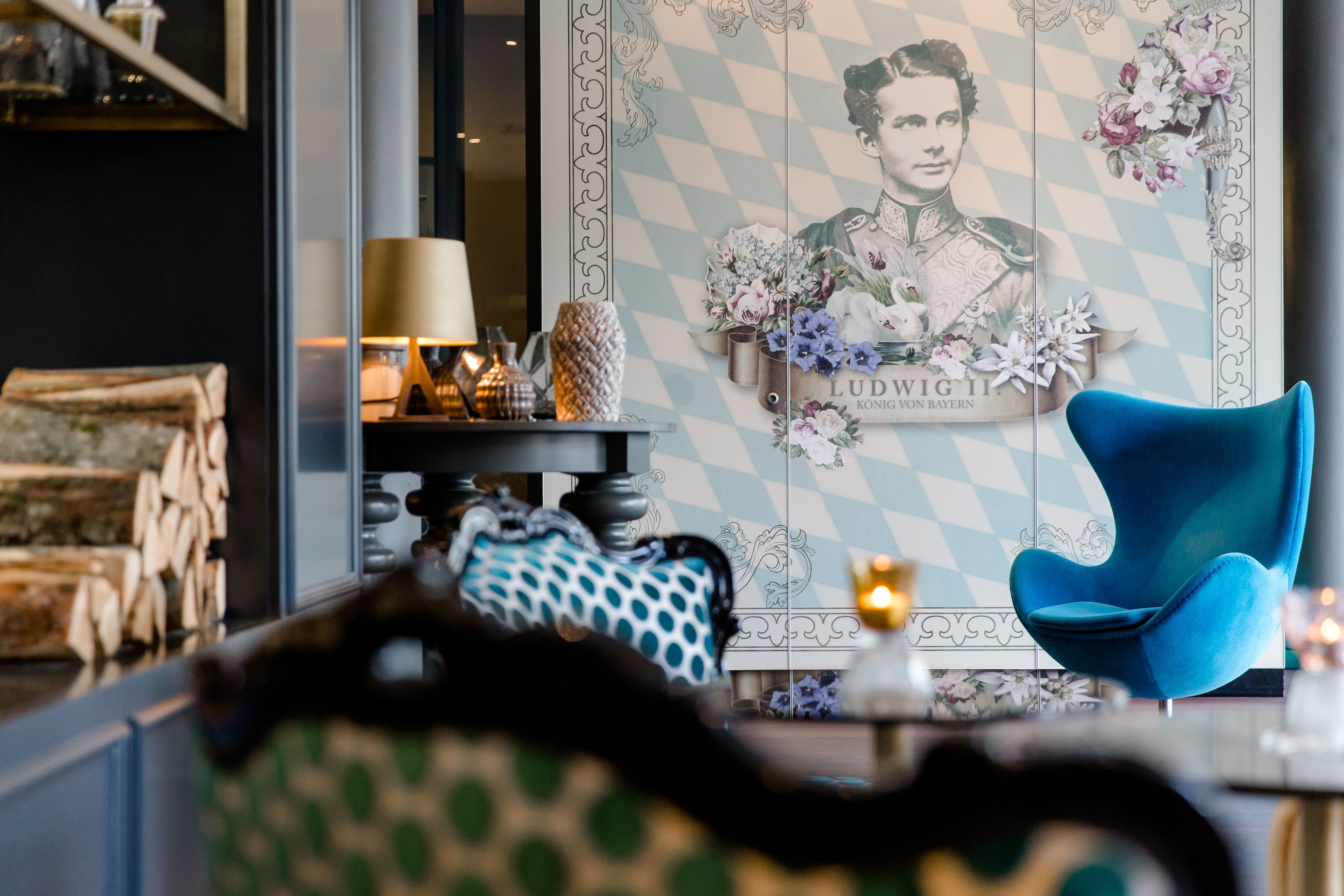 One Lounge mit blauem Designer-Sessel, gemusterten Sitzmöbeln und Wandbild von König Ludwig II.