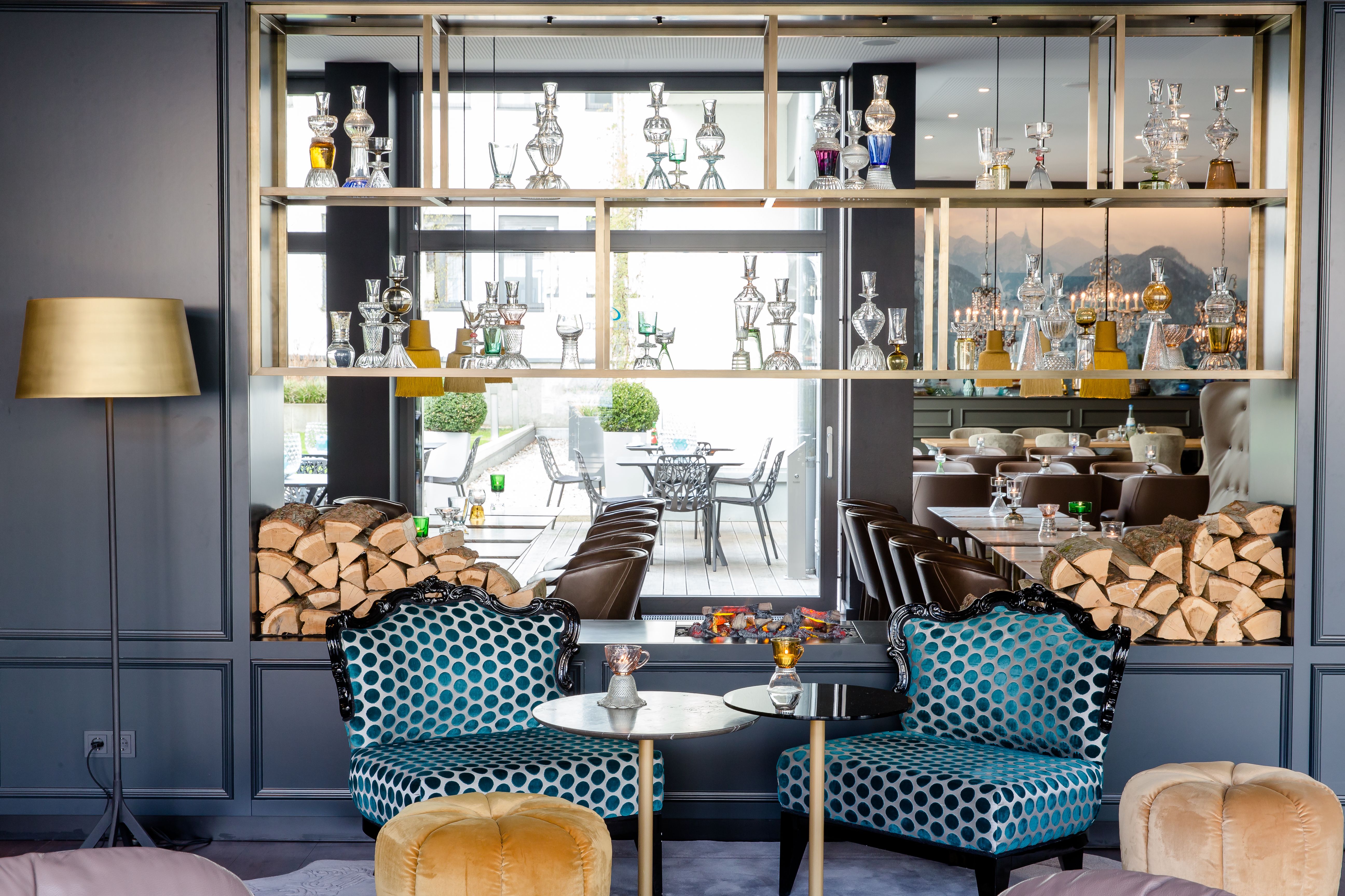 Motel One Lounge mit zwei gepolsterten Sesseln, kleinem Tisch und moderner Dekoration aus gläsernen Karaffen. Durch große Fenster im Hintergrund Blick auf Terrasse mit Sitzgelegenheiten und Bergpanorama. Stapel aus Feuerholz flankieren den Bereich.