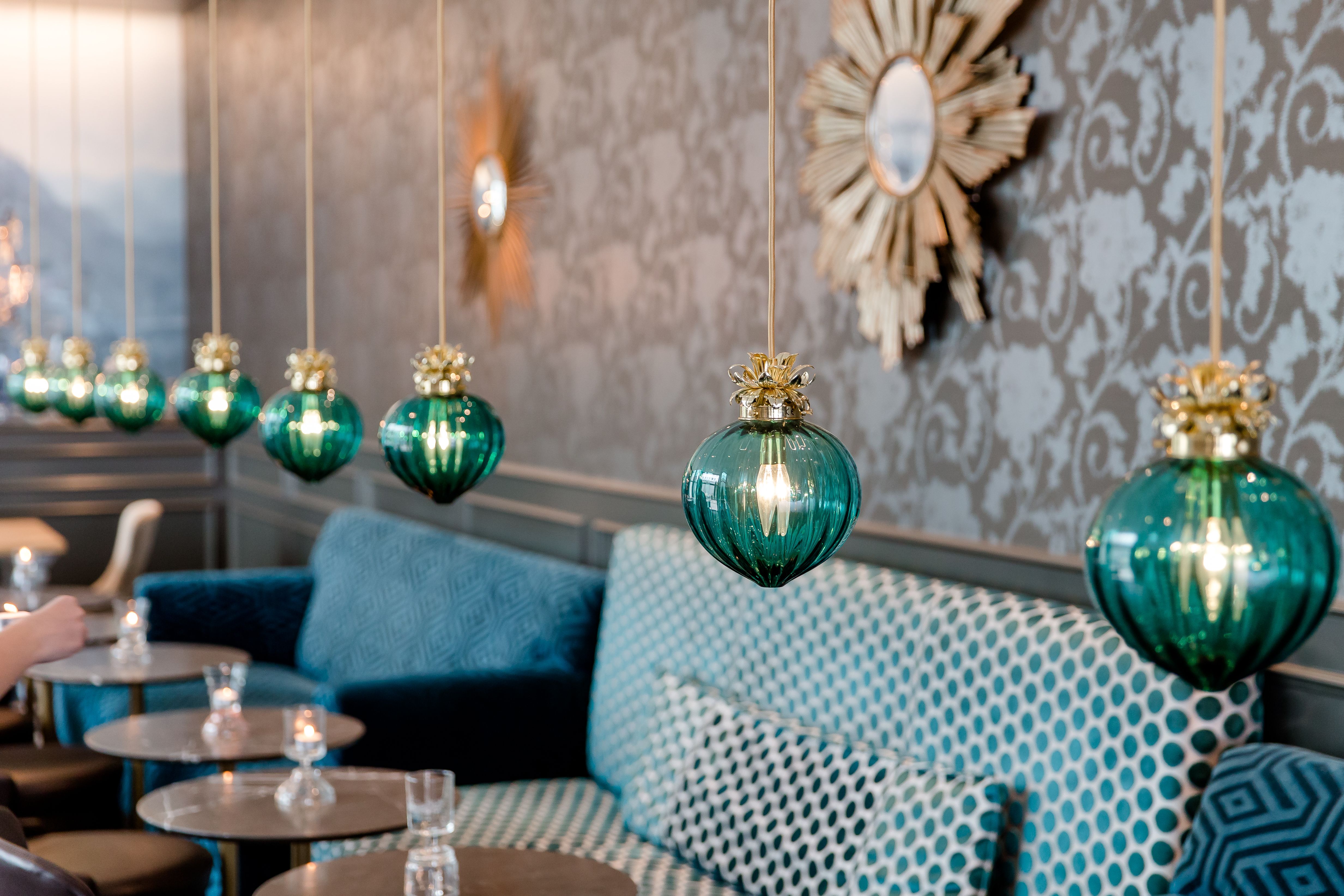 Stimmungsvolle Hotelbar mit eleganten, grünen Hängelampen, gepolsterten Sitzbänken in Musterstoffen und stilvoller Wanddekoration.