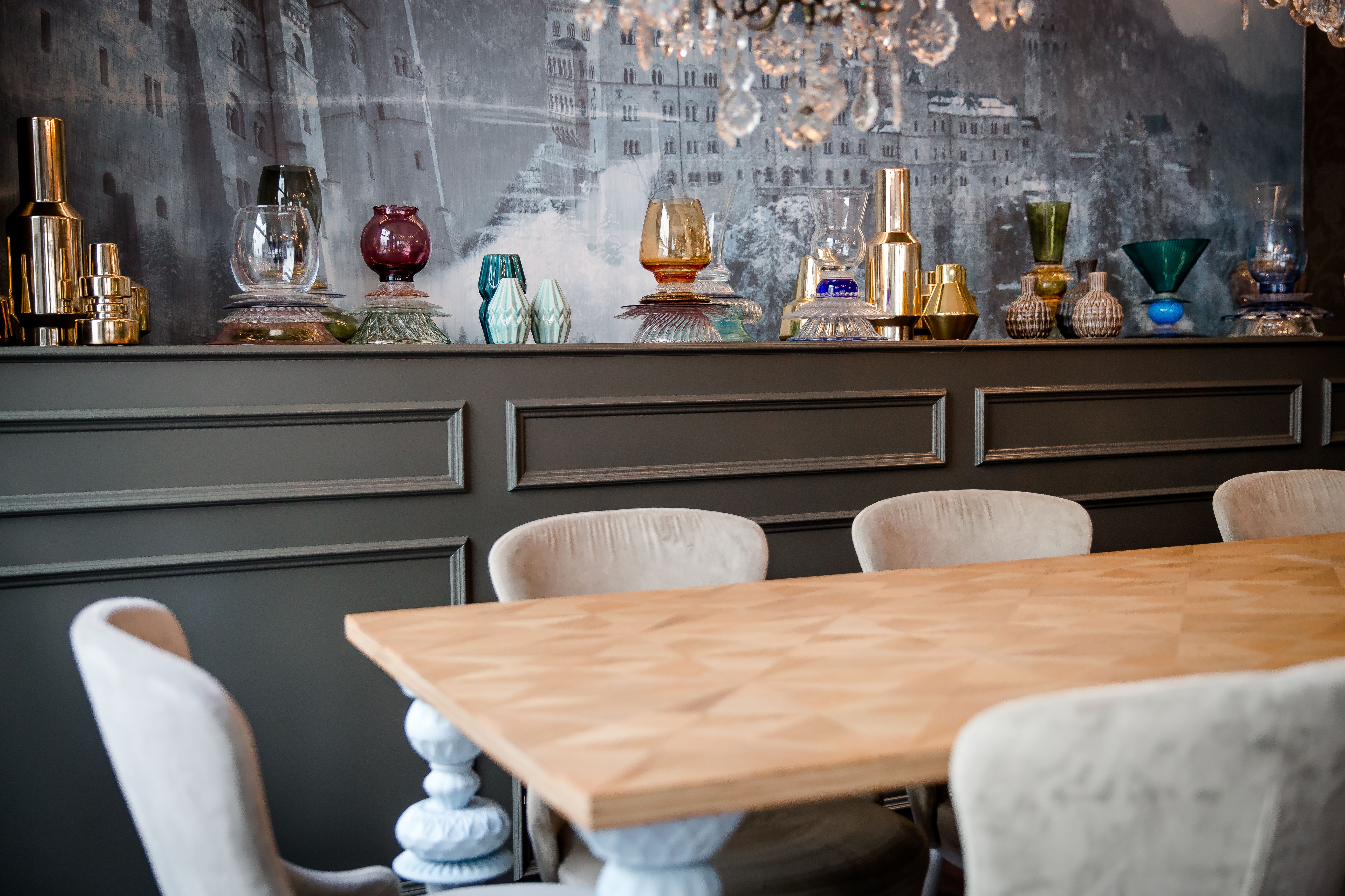 Workbench mit hellem Holztisch und bequemen, gepolsterten Stühlen. Im Hintergrund eine Wand mit dekorativem Sideboard, darauf bunte Glasobjekte und goldfarbene Accessoires.