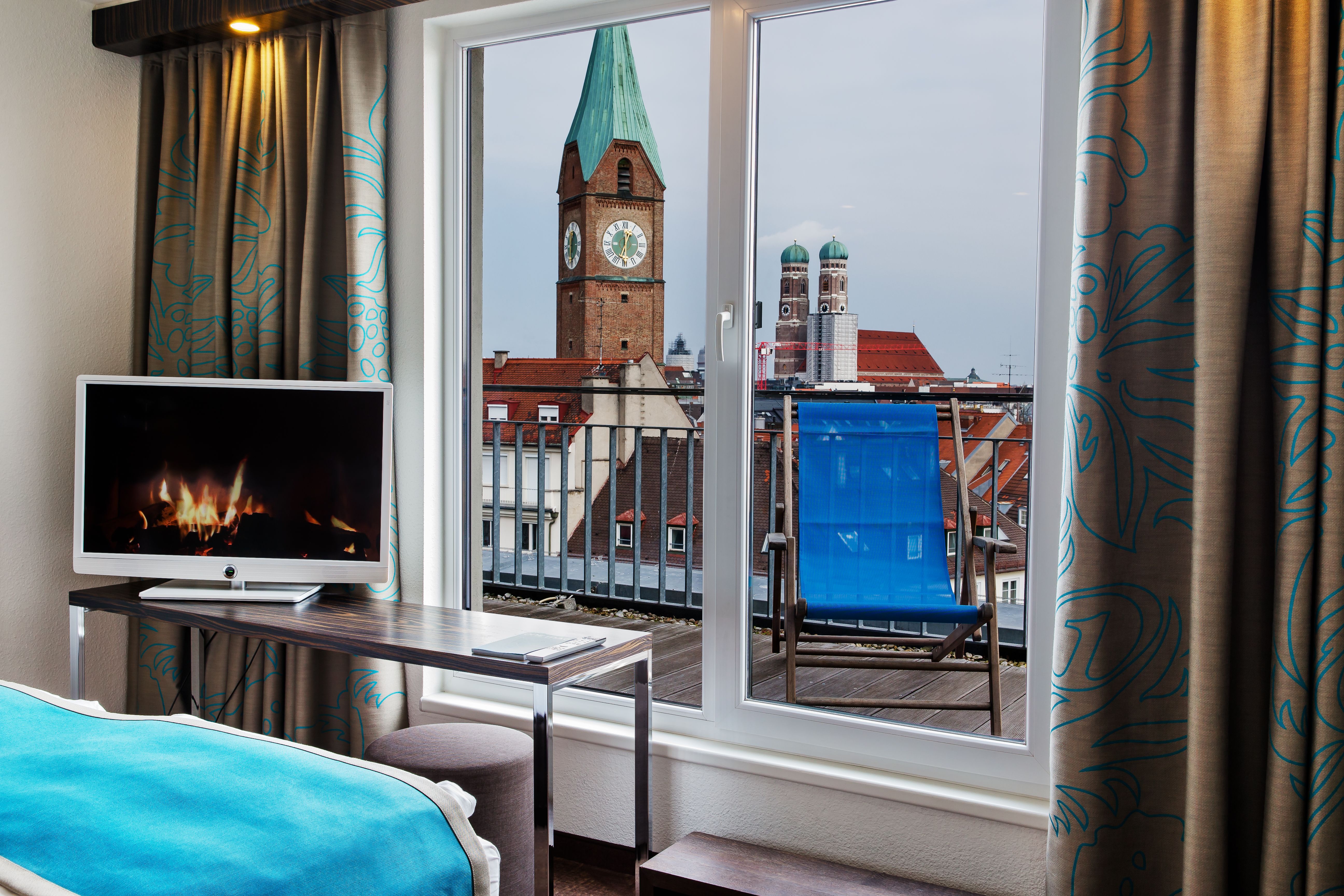Blick aus einem Hotelzimmer mit gemütlichem Bett und Fernseher auf Balkon mit Liegestuhl und die historischen Kirchtürme der Stadt.