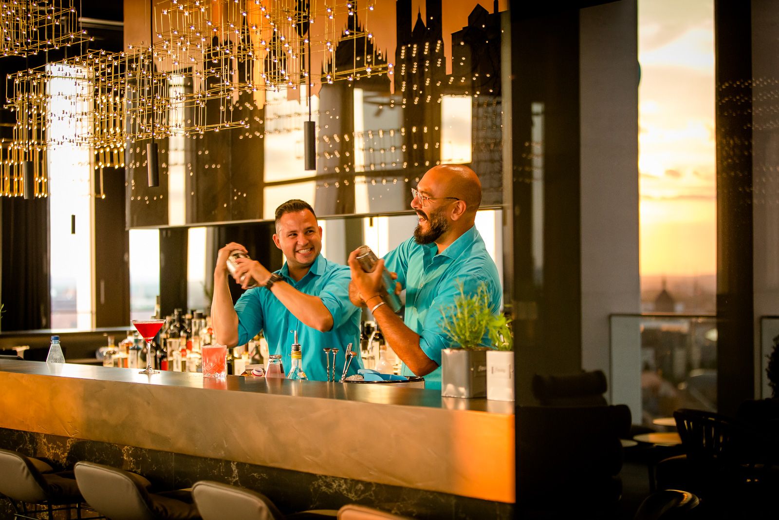 Zwei Barkeeper in blauen Hemden mixen Cocktails hinter einer stilvollen Bar bei Sonnenuntergang, umgeben von warmem Licht und moderner Einrichtung.