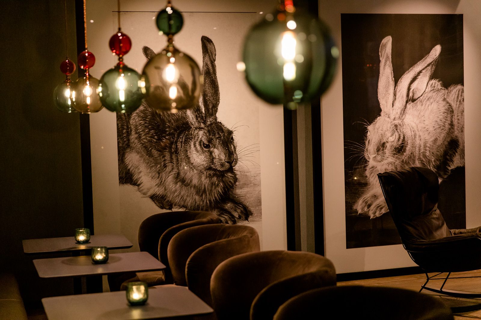 Aufnahmen eines stilvoll eingerichteten Raums mit weichen Sitzgelegenheiten, beleuchtet von warmen, hängenden Glühlampen. An der Wand sind große, detailreiche Schwarz-Weiß-Zeichnungen von Hasen zu sehen