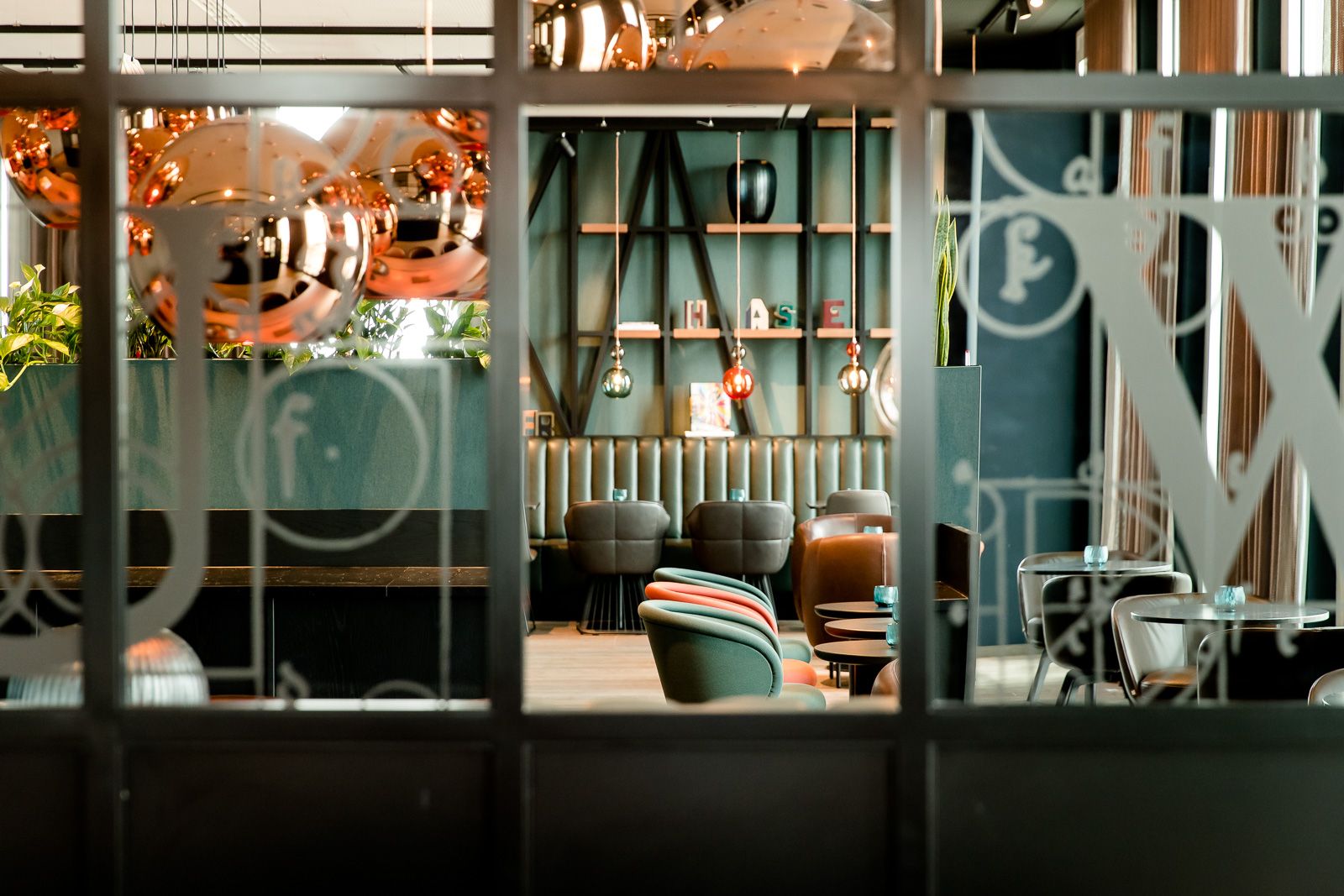 Blick durch Glastüren in eine moderne Motel One Hotel-Lounge mit gemütlichen Sesseln, dekorativen Kupferlampen und stilvoller Wandgestaltung