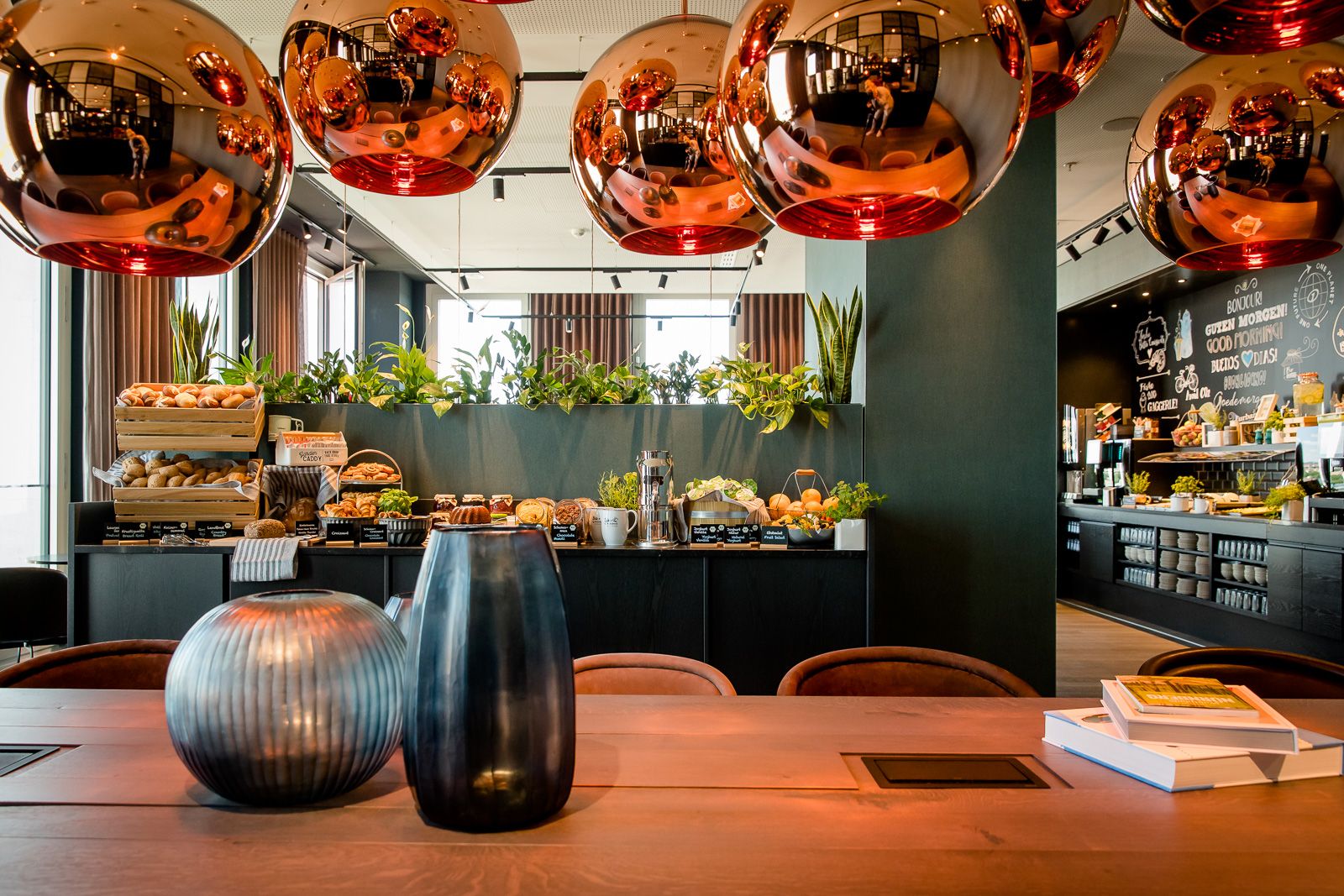 Motel One Frühstücksbereich mit einer Vielfalt an frischem Brot, Gebäck und Aufstrichen, umgeben von stilvollen Kupferlampen und grünen Pflanzen,