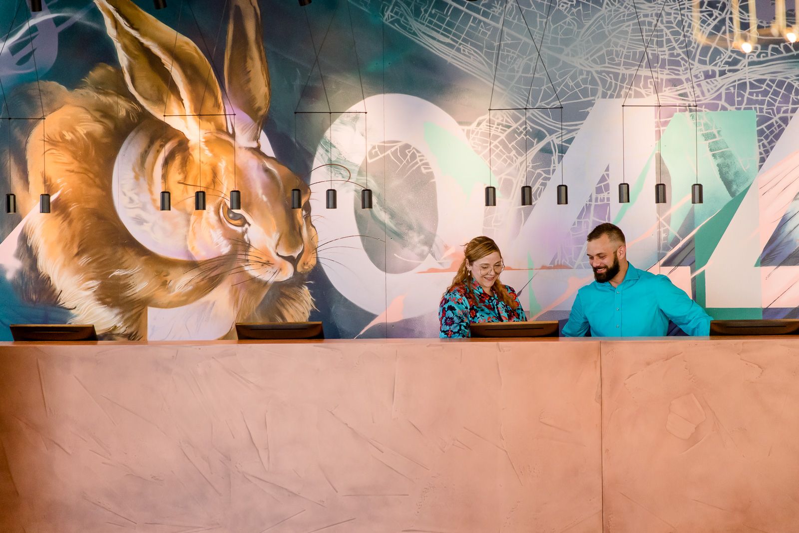 Zwei freundliche Empfangsmitarbeitende stehen hinter dem Empfangstresen vor einer farbenfrohen Wand mit großem Hasenmotiv und städtischem Kartendesign,.