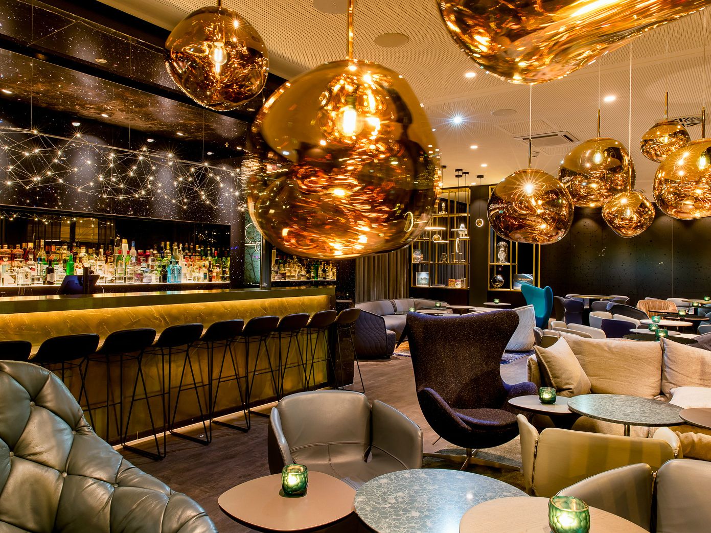Motel One Hotelbar mit warmem Licht, modernen Sitzgelegenheiten und stilvollen goldfarbenen Hängeleuchten.