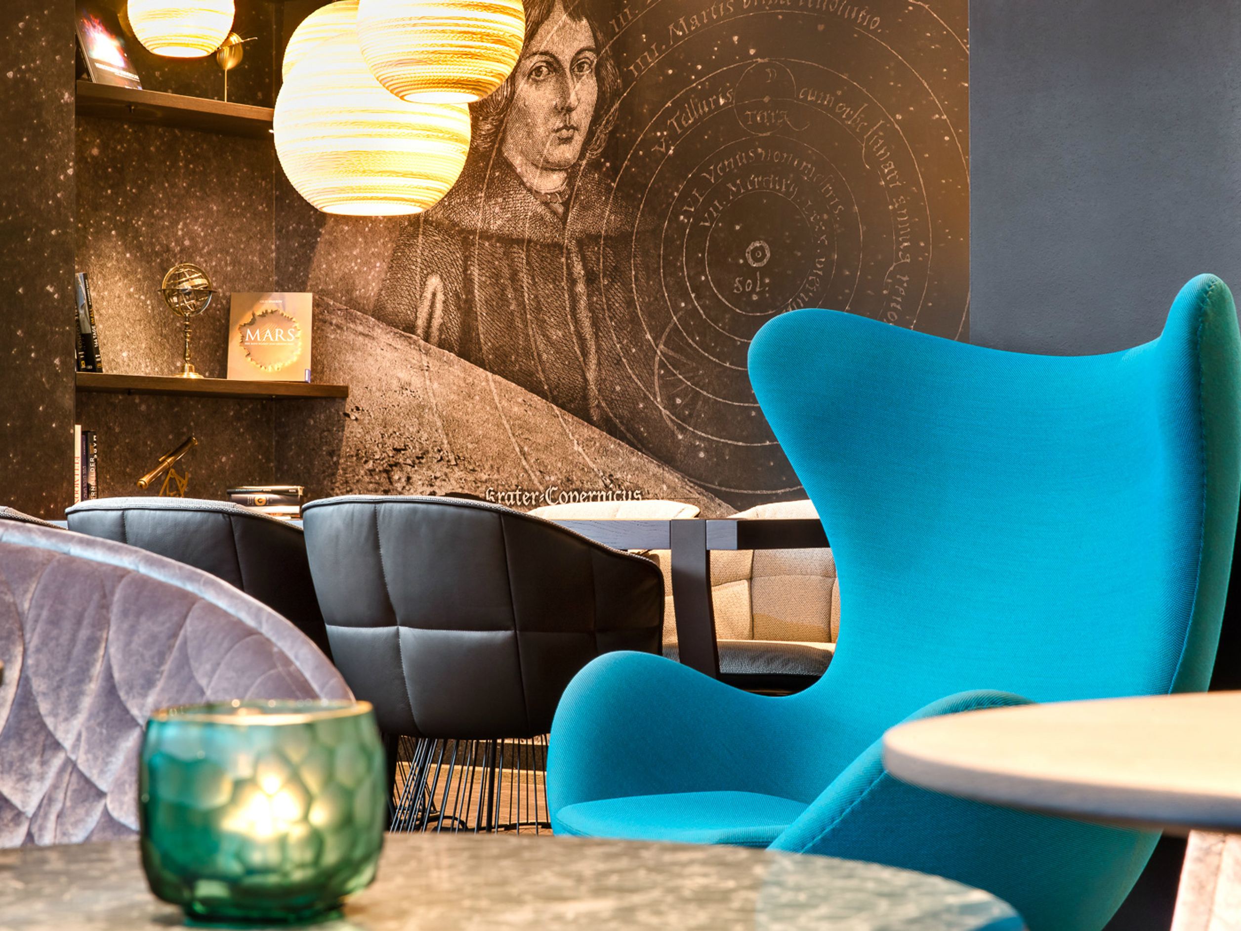 Motel One Lounge mit blauem Sessel, schwarzen Polsterstühlen und dekorativer Wandgrafik eines Astronomen