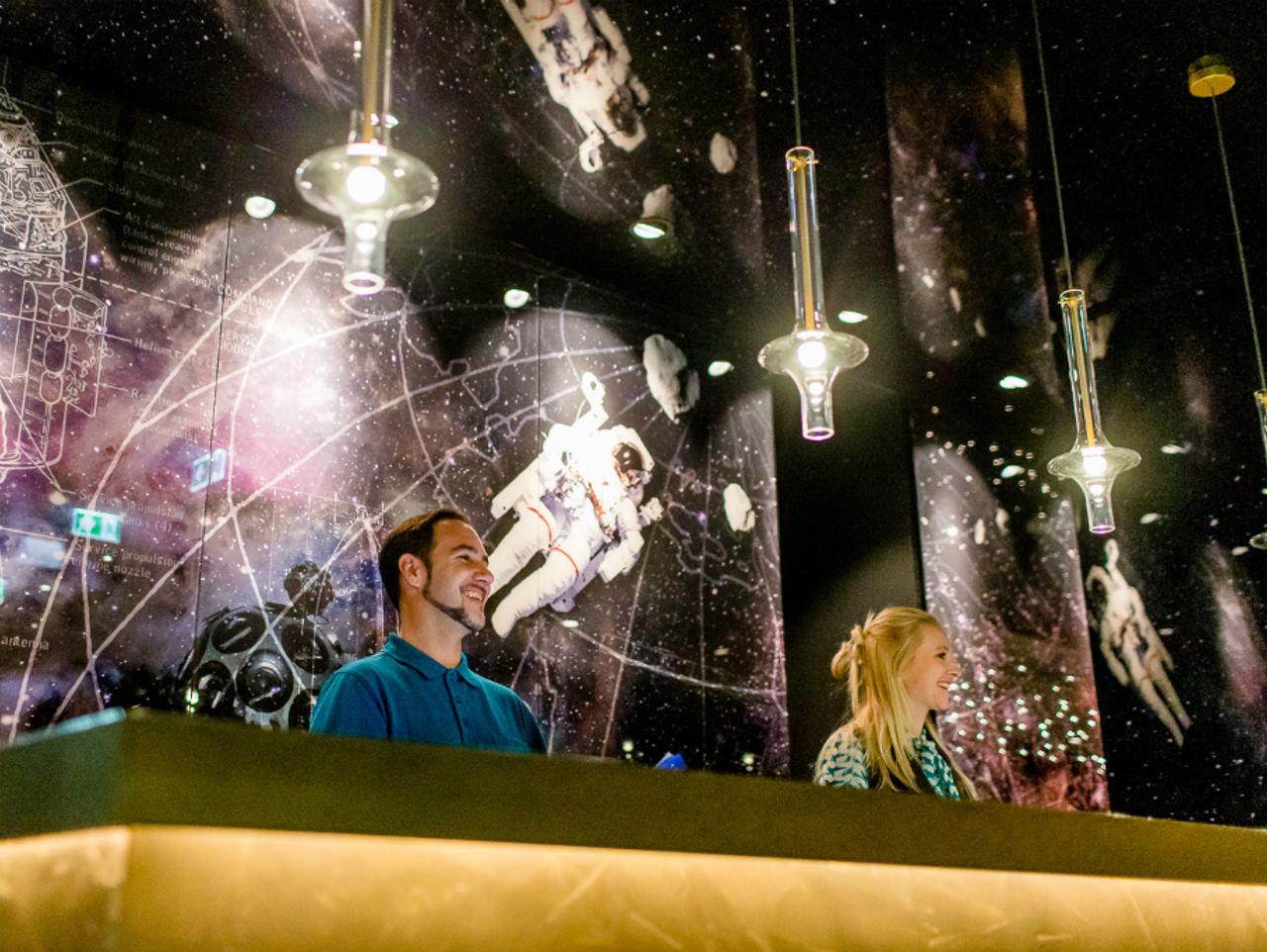 Zwei freundliche Mitarbeiter an der Rezeption eines Hotels mit einer modernen, weltrauminspirierten Wandgestaltung, die Astronauten und Galaxien zeigt