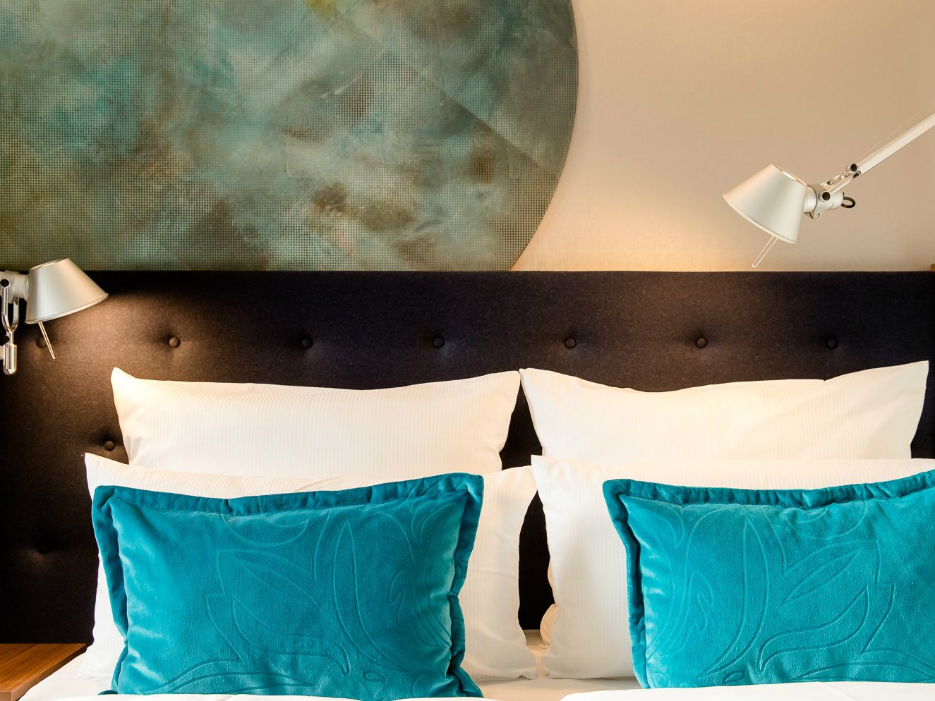 Gemütliches Motel One Doppelbett mit dunkel gepolstertem Kopfteil, weißen Kissen und türkisfarbenen Dekokissen, beleuchtet von zwei verstellbaren Leselampen, vor einer Wand mit großem, rundem Wanddekor in beruhigenden Grüntönen.