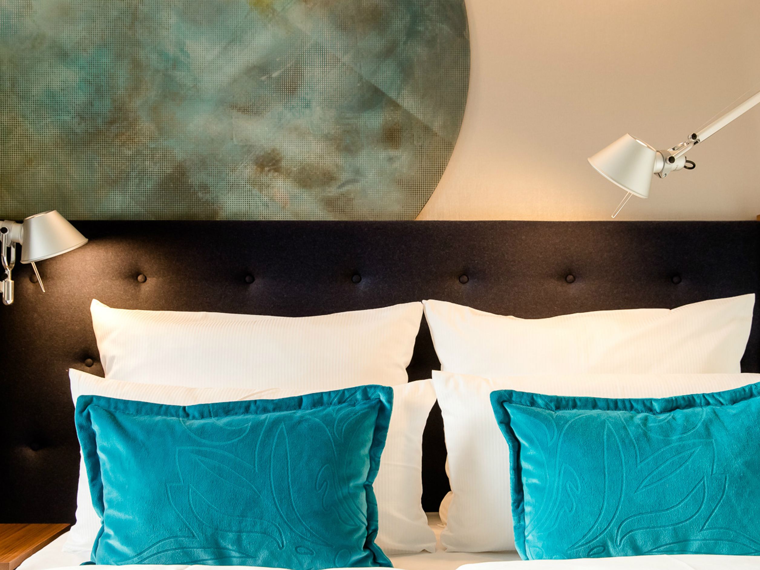 Gemütliches Motel One Doppelbett mit dunkel gepolstertem Kopfteil, weißen Kissen und türkisfarbenen Dekokissen, beleuchtet von zwei verstellbaren Leselampen, vor einer Wand mit großem, rundem Wanddekor in beruhigenden Grüntönen.