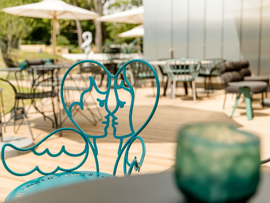 Drahtkunst in Form eines Paares, das sich küsst, als Stuhlrücken gestaltet. Im Hintergrund eine sonnige Außenterrasse mit Tischen, Stühlen und Sonnenschirmen