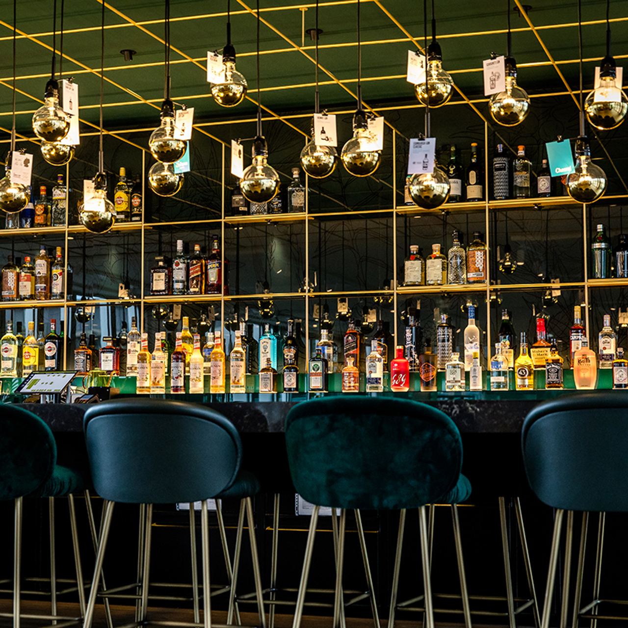 Motel One Hotelbar mit gemütlichen grünen Samthockern, beleuchtet von vielen dekorativen Glühlampen, die über einer gut sortierten Auswahl an Spirituosen und Getränken an einer eleganten schwarzen Theke schweben.