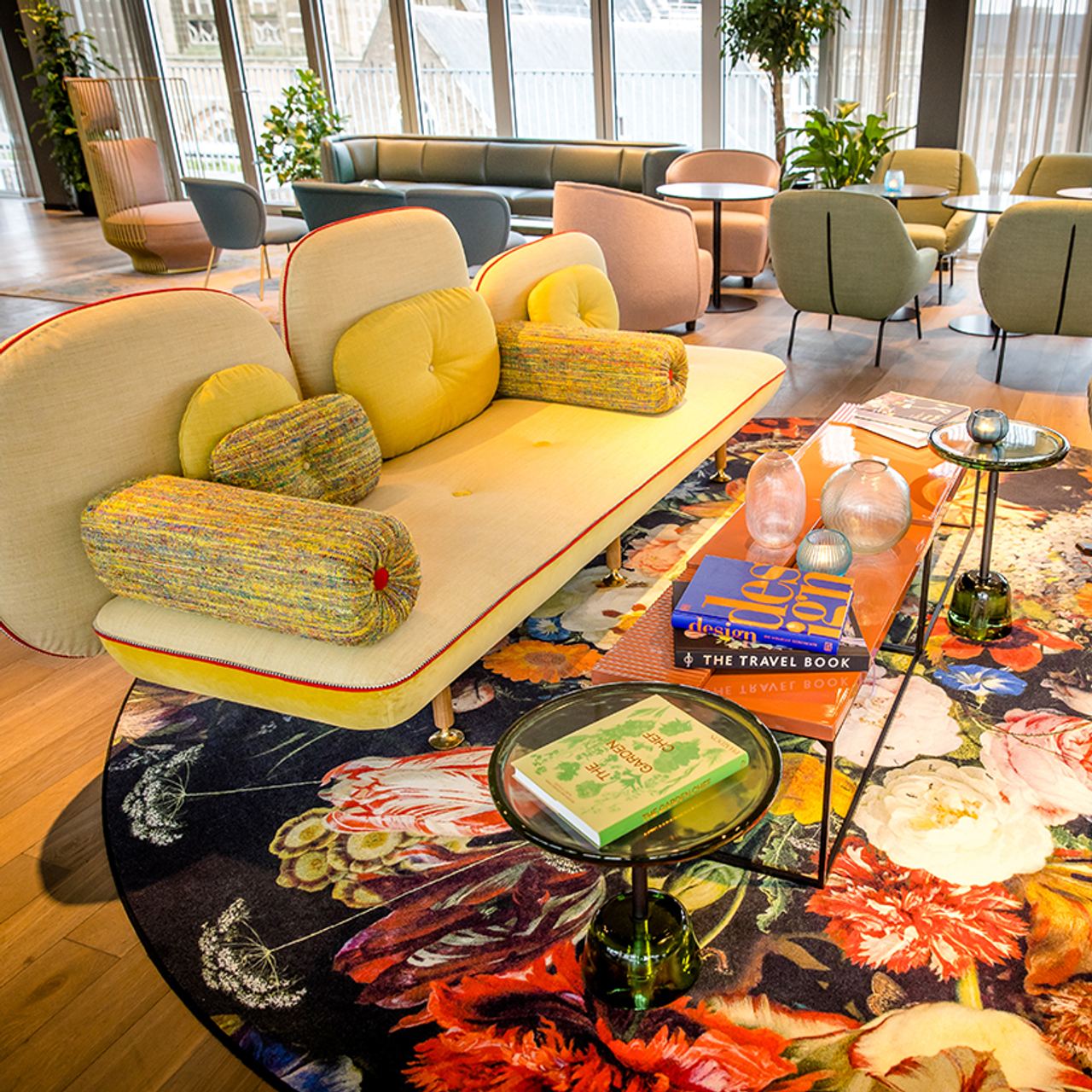 Motel One Lounge mit farbenfrohem Sofa und dekorativem Teppich in lebendigem Blumenmuster. Gläserne Beistelltische mit Büchern und stilvollen Deko-Elementen, großer heller Raum mit Sitzgruppen und Pflanzen