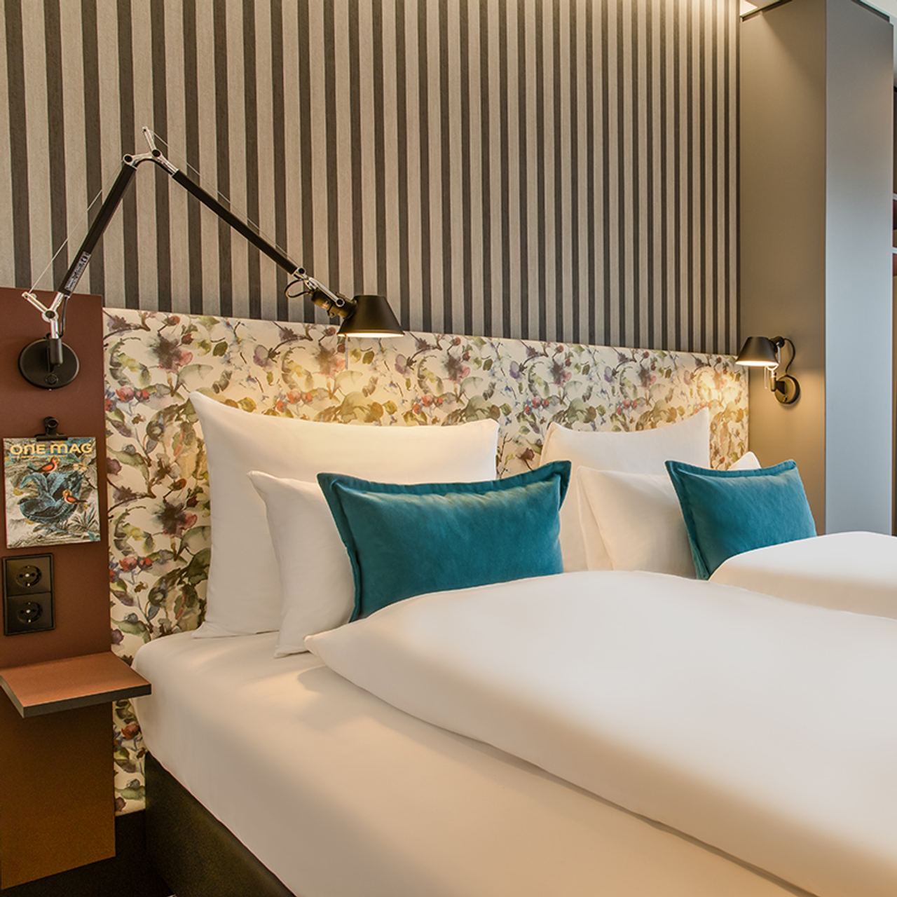 Motel One Doppelzimmer mit floralem Kopfteil, gestreifter Tapete, weißen Bettwäsche und türkisen Akzentkissen, sanfter Beleuchtung und schlichtem Nachttisch.