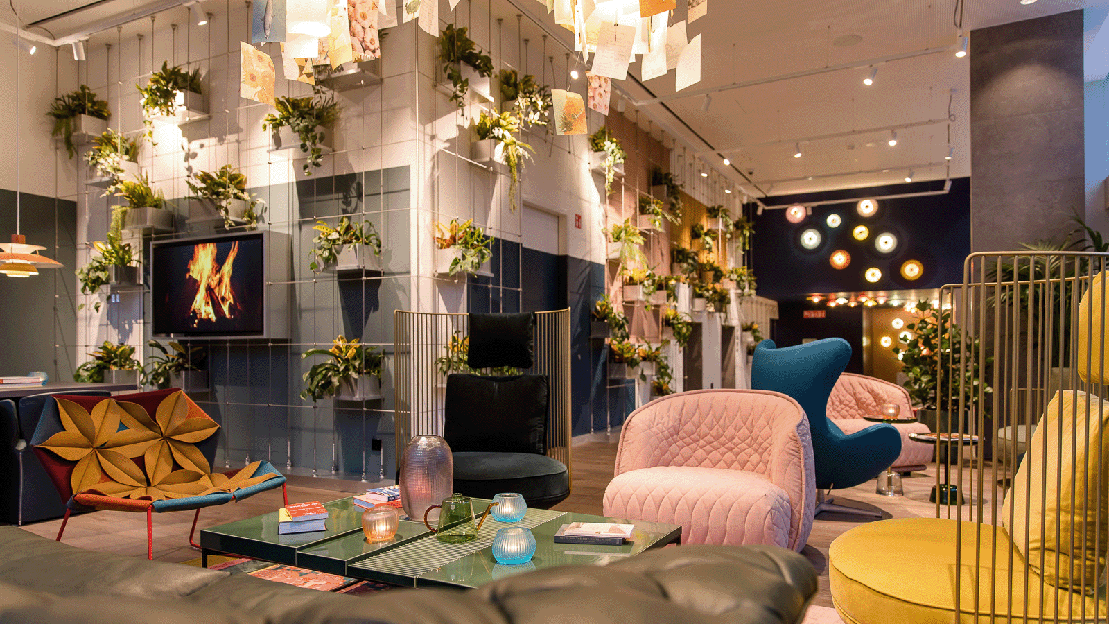 Motel One Hotel-Loungebereich mit modernen, farbenfrohen Sesseln, grünen Pflanzen an der Wand und einem dekorativen Kamin, der Wärme und Entspannung ausstrahlt. 