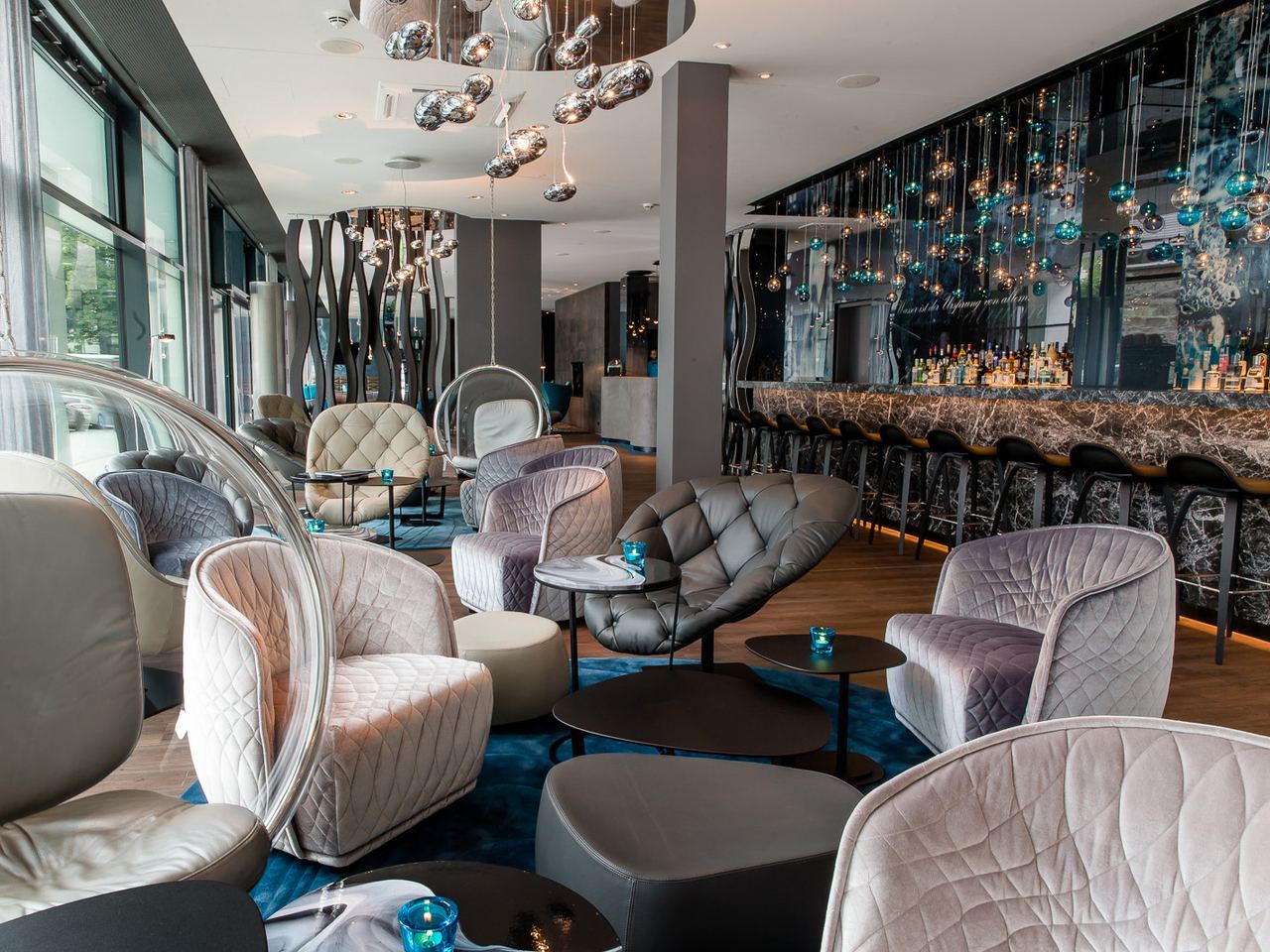 Motel One Hotelbar mit stilvollen, bequemen Sesseln und Sofas in gedeckten Farben, dekorativen Hängeleuchten und einer eleganten Bar mit breitem Getränkeangebot. Große Fenster sorgen für viel Tageslicht und Ausblick nach draußen.