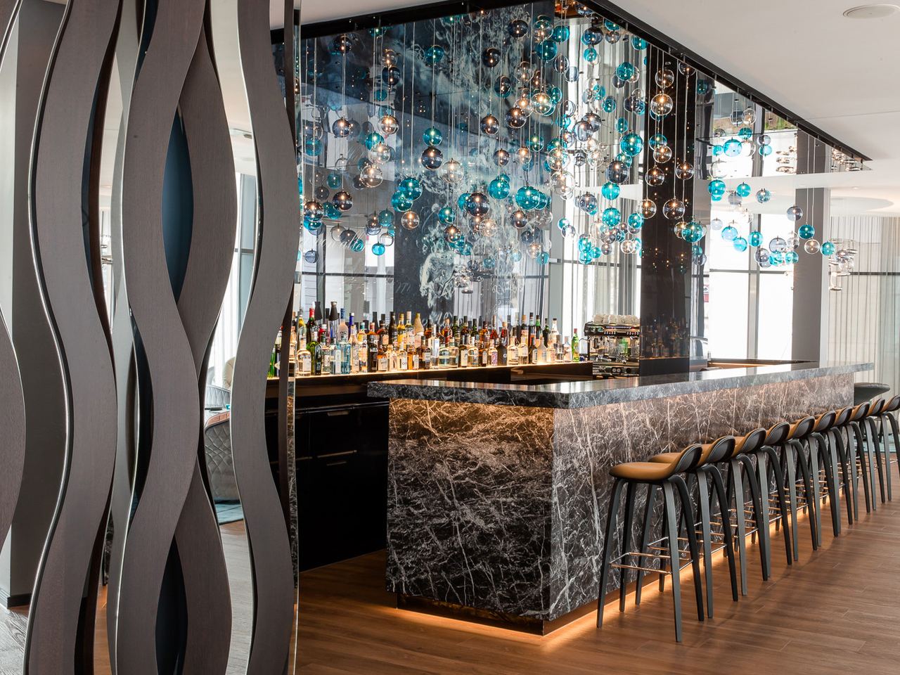 Motel One Hotelbar mit eleganter Marmor-Theke, stilvollen Barhockern und dekorativen, hängenden Glaskugeln in Blau- und Silber-Tönen