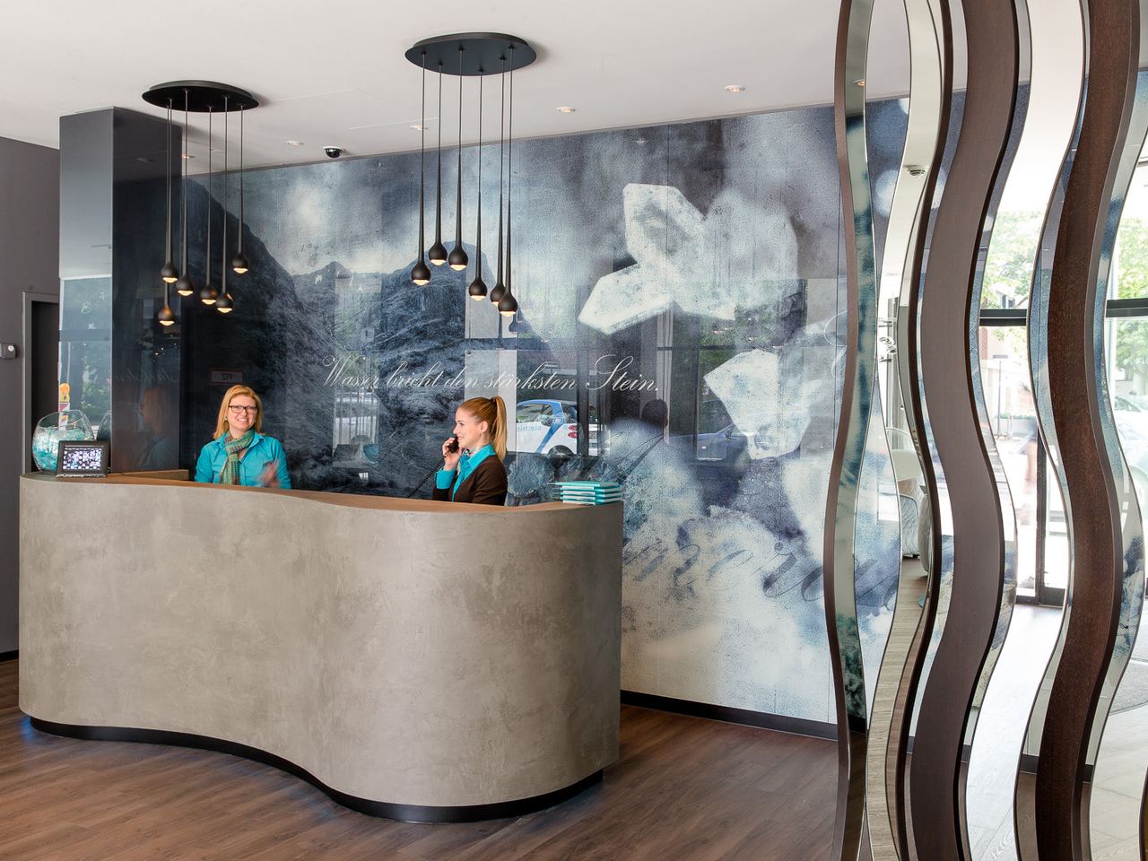 Rezeption mit moderner, klarer Gestaltung, zwei freundlichen Mitarbeiterinnen und sanfter Beleuchtung, gemütliche Atmosphäre für einen herzlichen Empfang im Hotel.