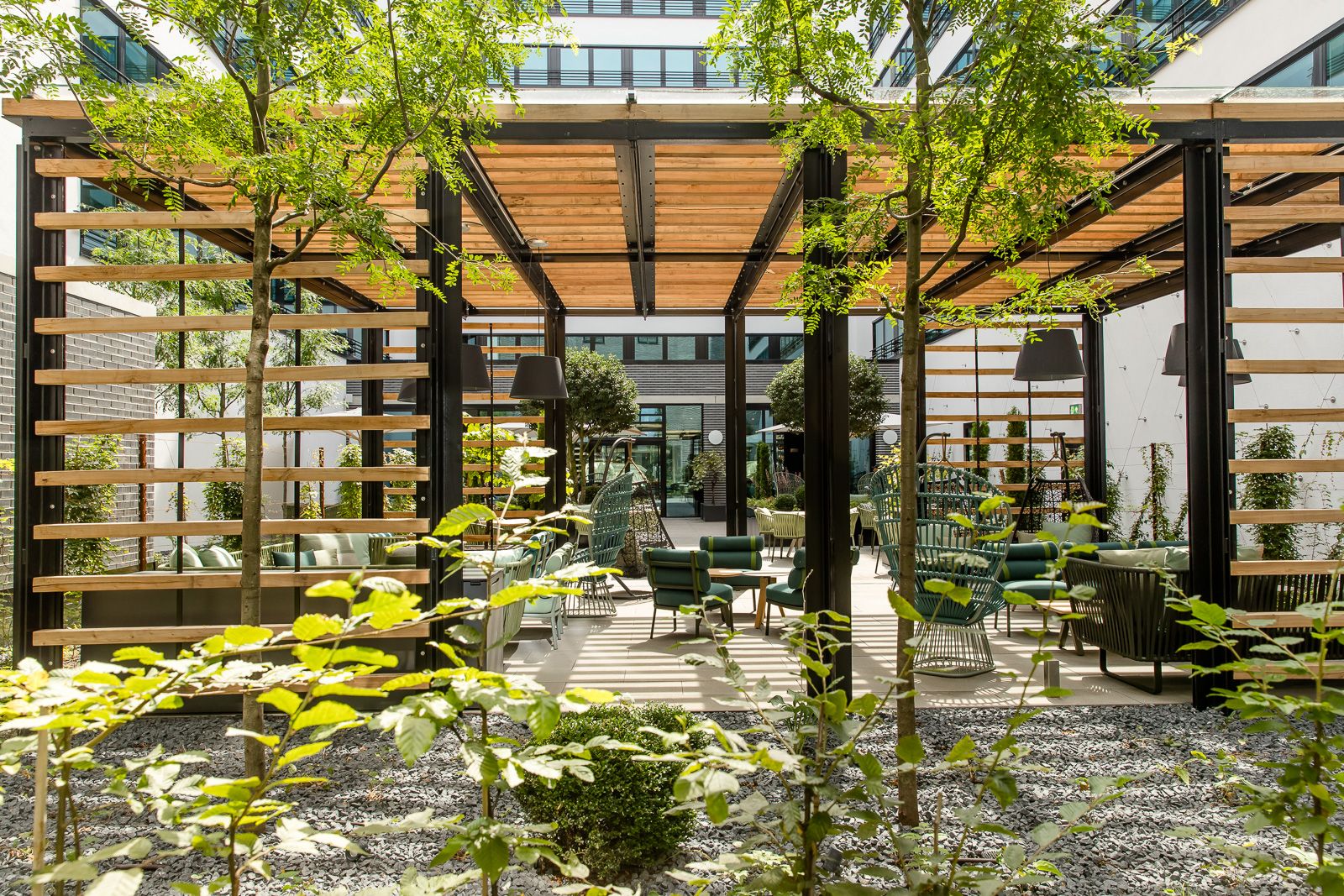 Lichtdurchfluteter Motel One Außenbereich mit grünen Pflanzen, gemütlichen Sitzmöbeln und Sonnenschutz. 