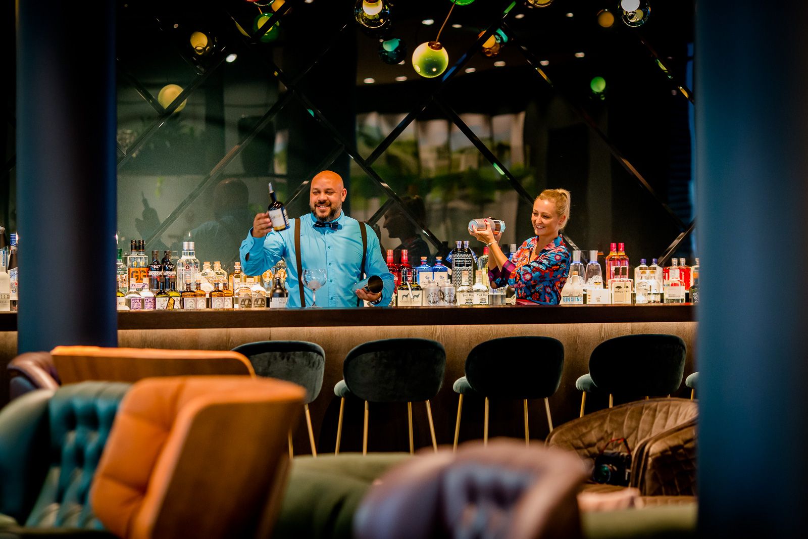 Zwei Barkeeper bereiten hinter einer stilvollen Bar Cocktails vor, umgeben von einer eleganten Atmosphäre mit bequemen Sitzgelegenheiten und Beleuchtung.