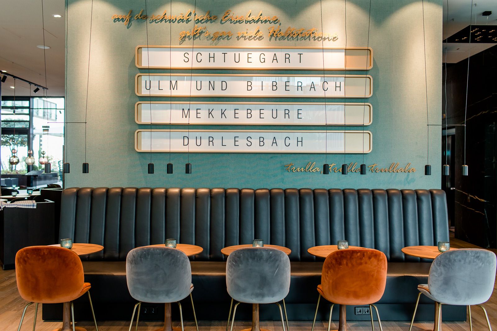 Wand mit stilvoller Beschriftung traditioneller Ortsnamen, darunter Sitzbank in dunklem Leder, daneben bunte samtige Stühle und runde Tische