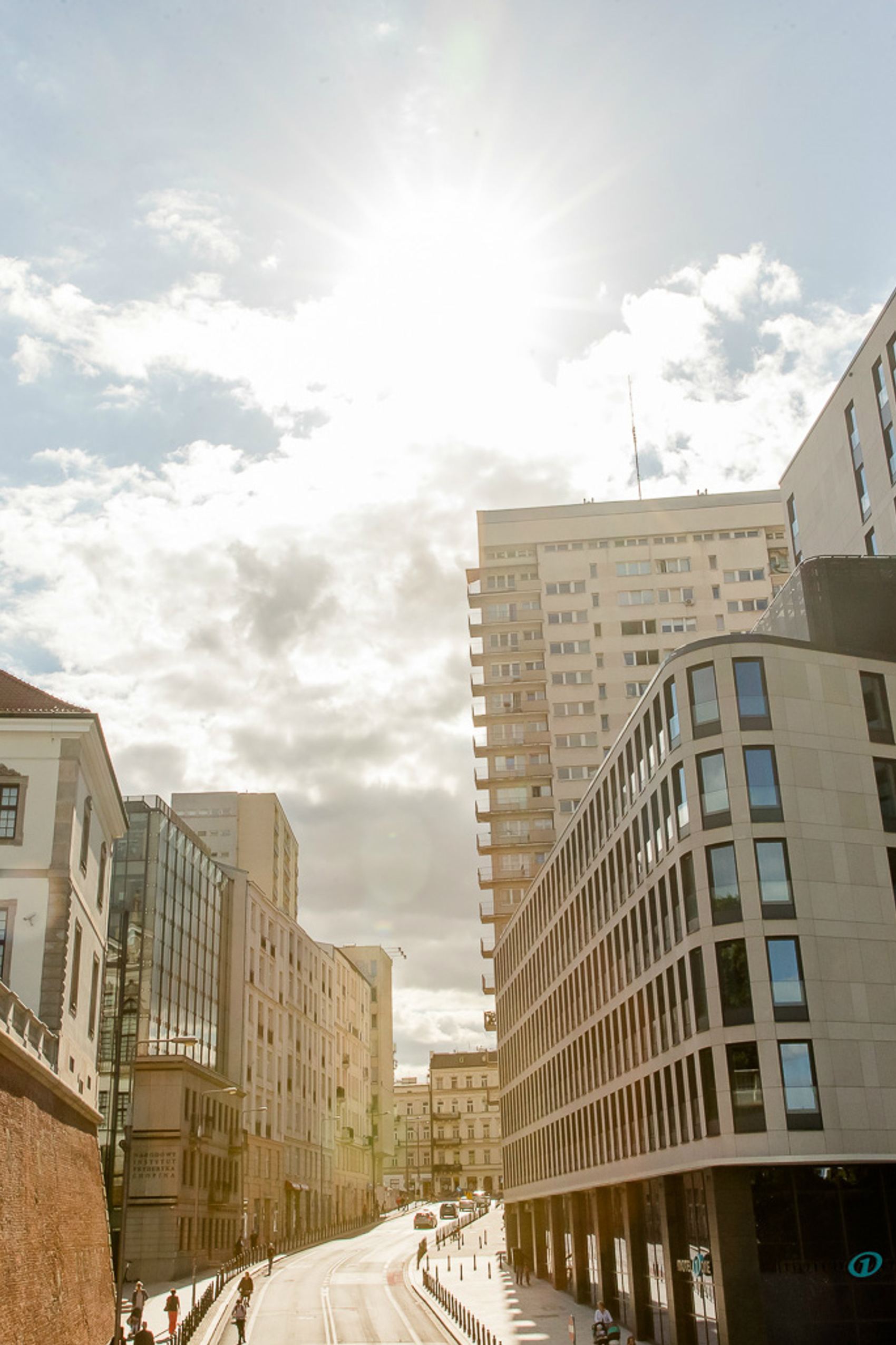 Straßenszene bei Sonnenschein mit moderner und historischer Architektur, lichtdurchfluteten Gebäuden und vereinzelten Menschen auf dem Bürgersteig.