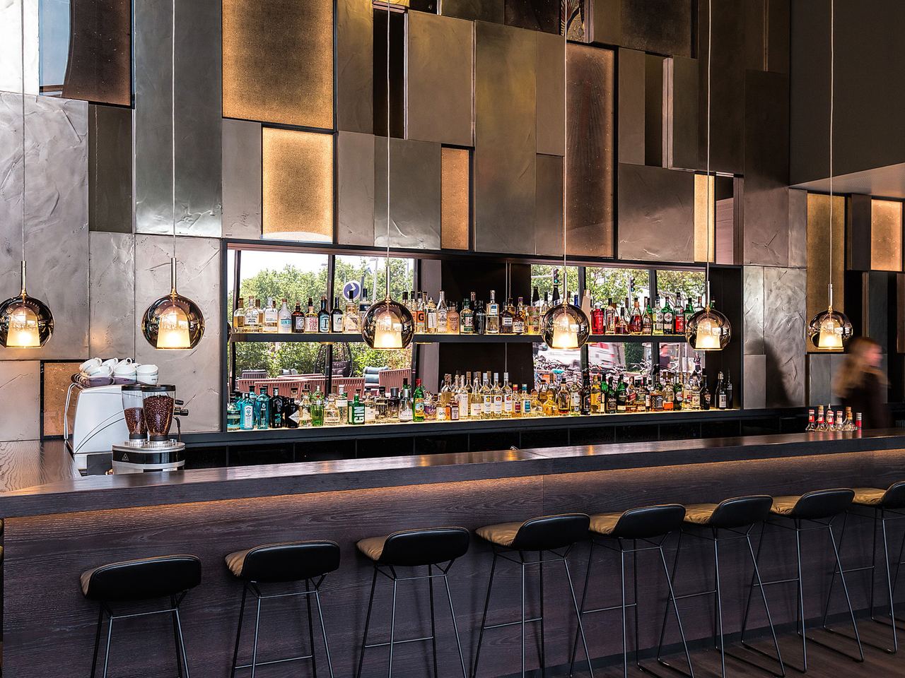 Motel One Hotelbar mit langem, dunklem Tresen und sechs schwarzen Barhockern, beleuchtet von modernen Hängeleuchten. Im Hintergrund sind vielfältige Spirituosen und große Fenster mit Außenblick zu sehen.