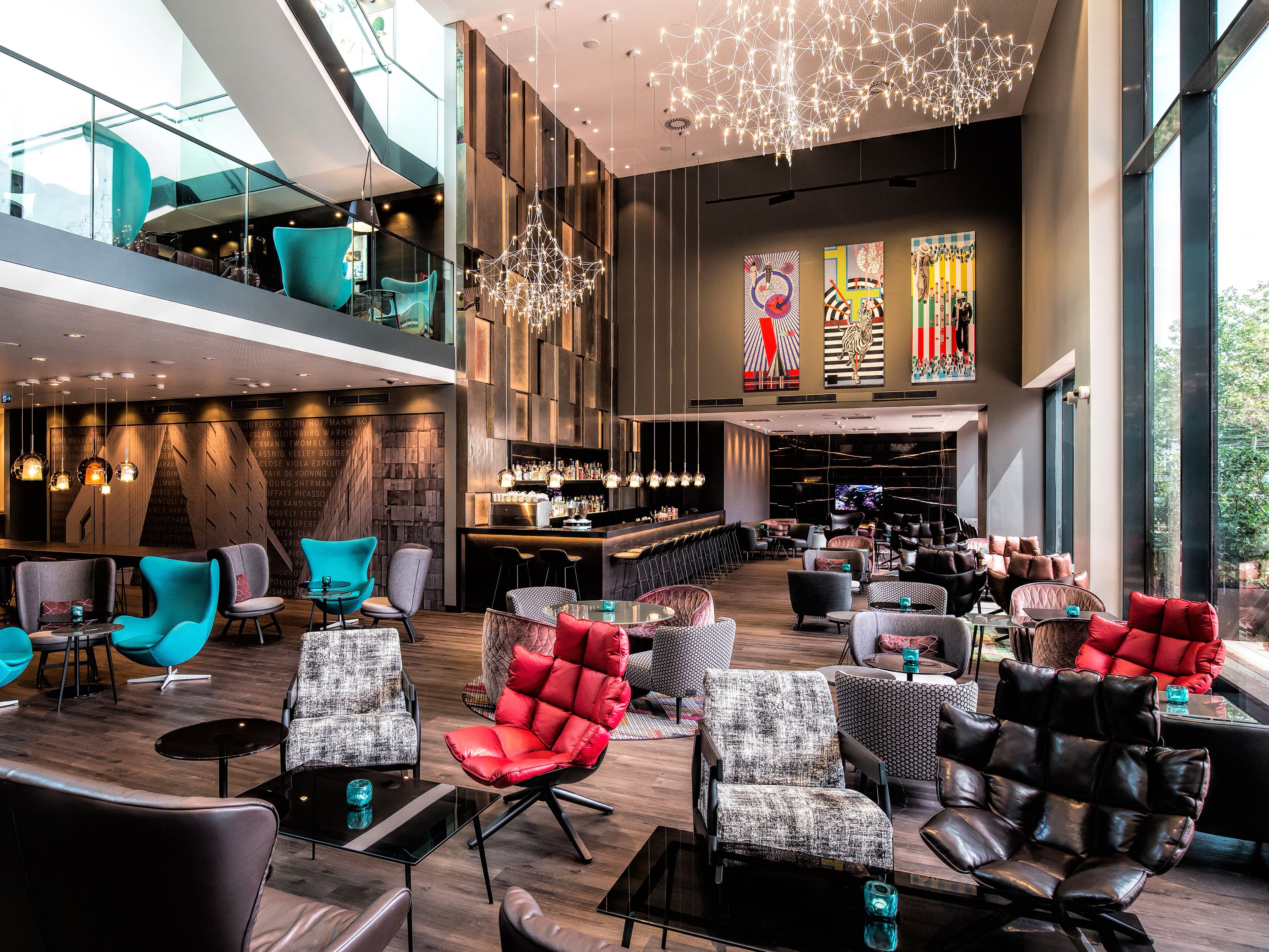 Motel One Hotel-Loungebereich mit hohen Decken, stilvollen Sesseln in verschiedenen Farben, großen Fensterfronten für viel Tageslicht und kunstvollen Deckenleuchten.