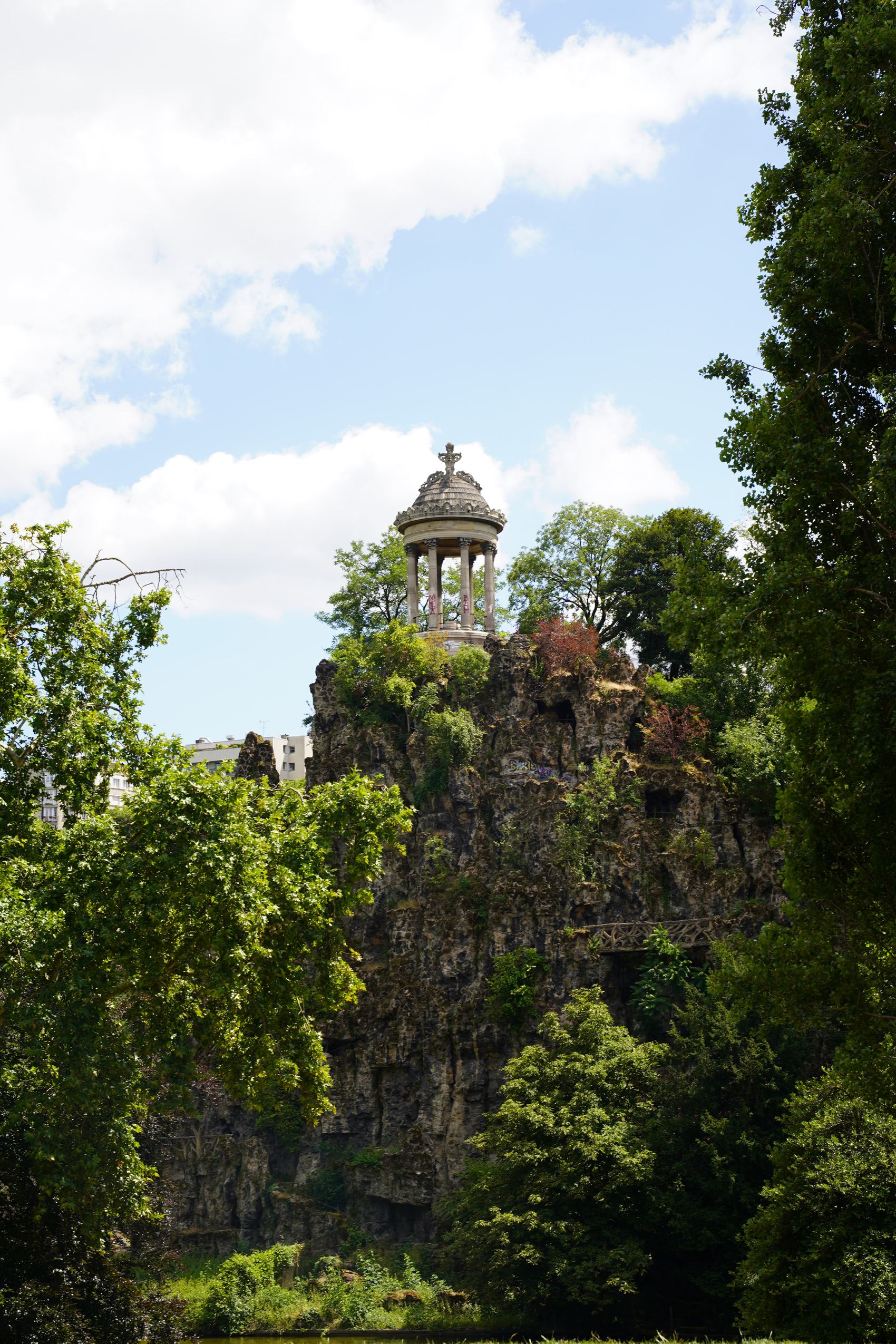Ein historisches Pavillon-ähnliches Bauwerk auf einer felsigen Anhöhe, umgeben von dichtem grünem Laub, unter blauem Himmel mit vereinzelten Wolken.