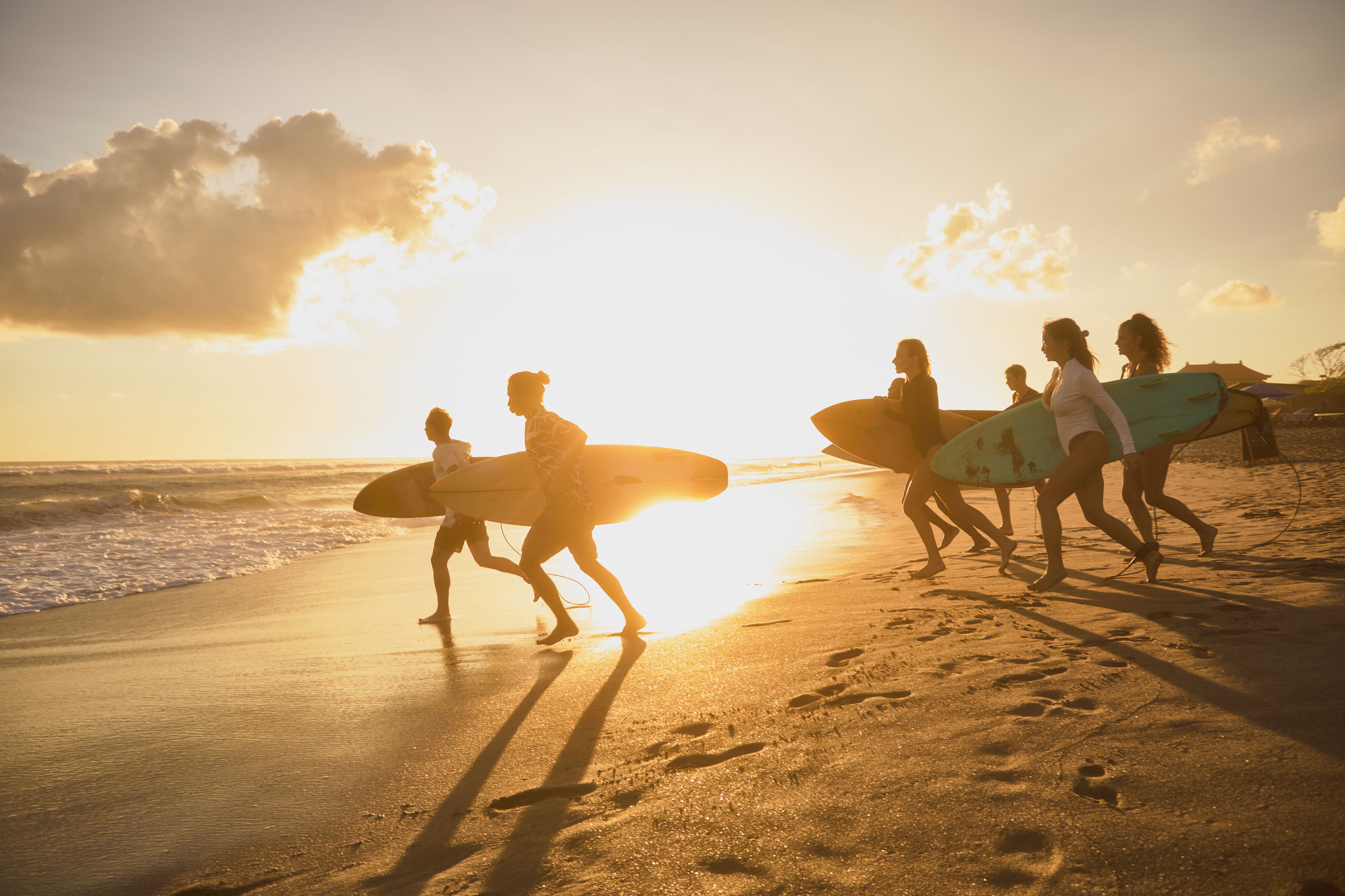 Fünf Personen tragen Surfbretter und laufen bei Sonnenuntergang am Strand Richtung Meer, bereit zum Surfen. Die warme Lichtstimmung vermittelt Abenteuerlust und Freiheit.