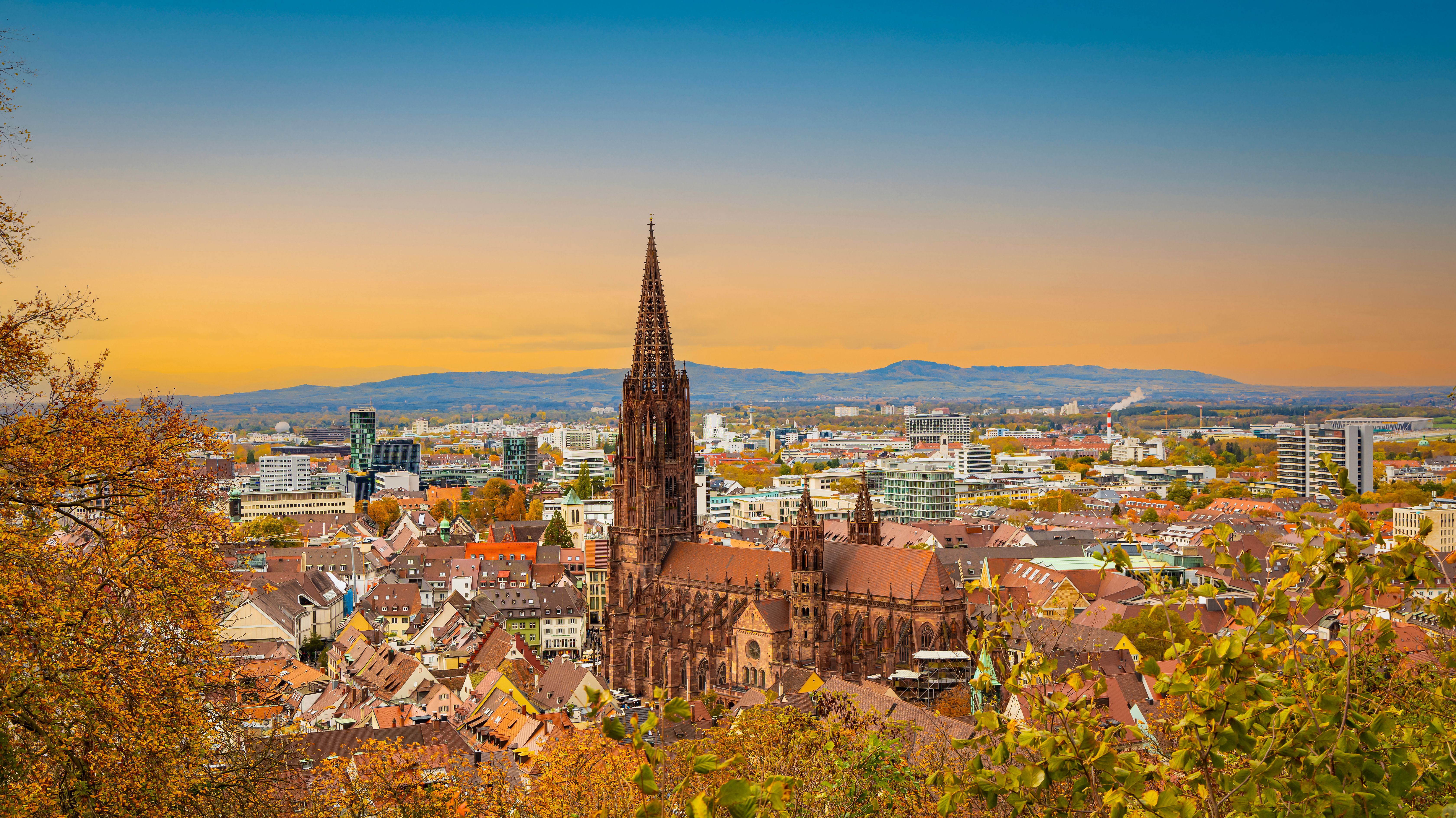 Blick über eine Stadt mit historischen Gebäuden und einer markanten gotischen Kathedrale im Zentrum, umgeben von herbstlich gefärbtem Laub und einer sanften Abenddämmerung am Himmel.