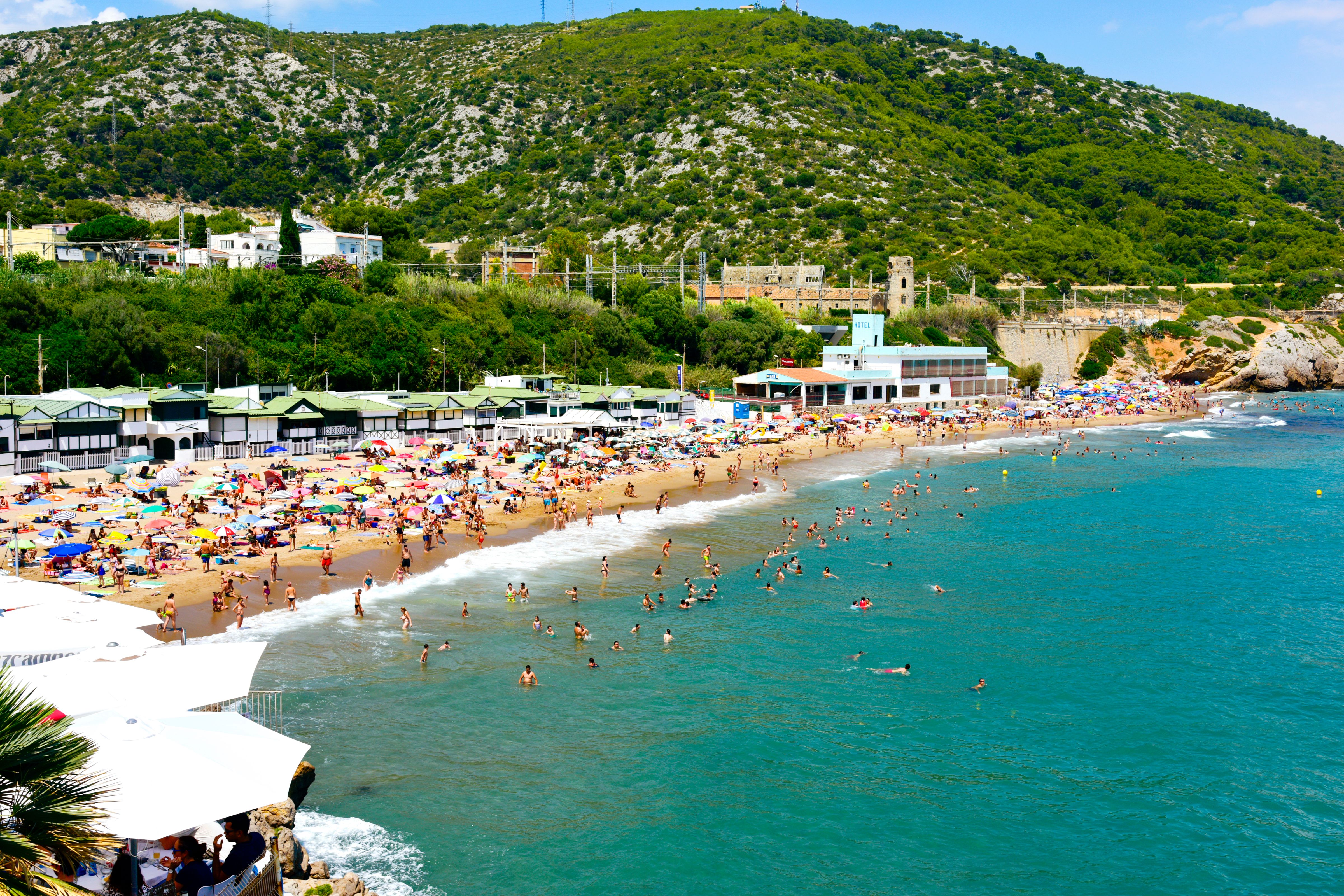 Strand mit vielen Sonnenschirmen und Badegästen im Wasser und am Sand, davor Hügel mit grüner Vegetation, vermitteln lebhafte Urlaubsatmosphäre an der Küste.