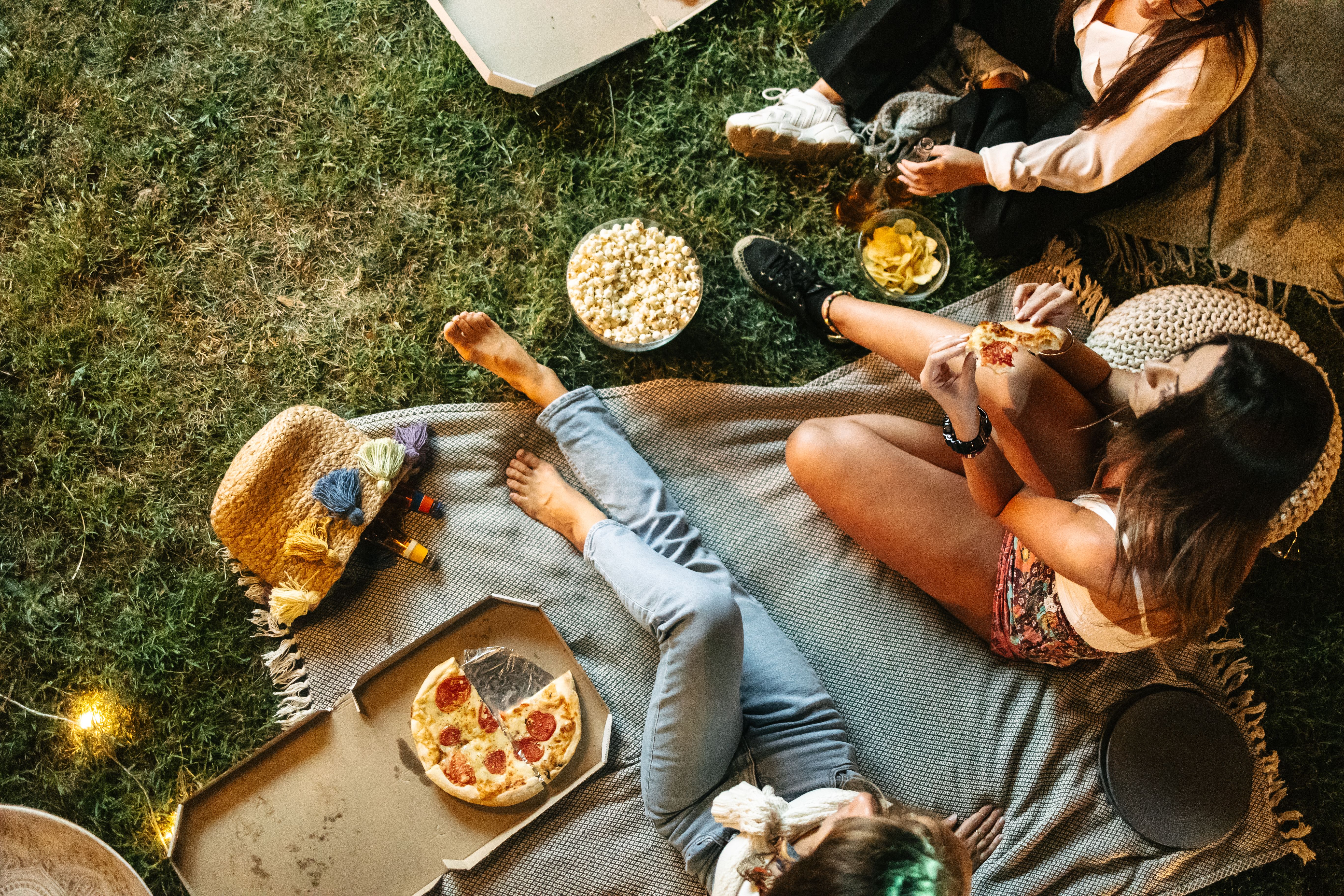 Drei Frauen sitzen gemütlich auf einer Decke im Gras, essen Pizza und Snacks, umgeben von warmem, stimmungsvollem Licht, und genießen einen entspannten, geselligen Abend im Freien.