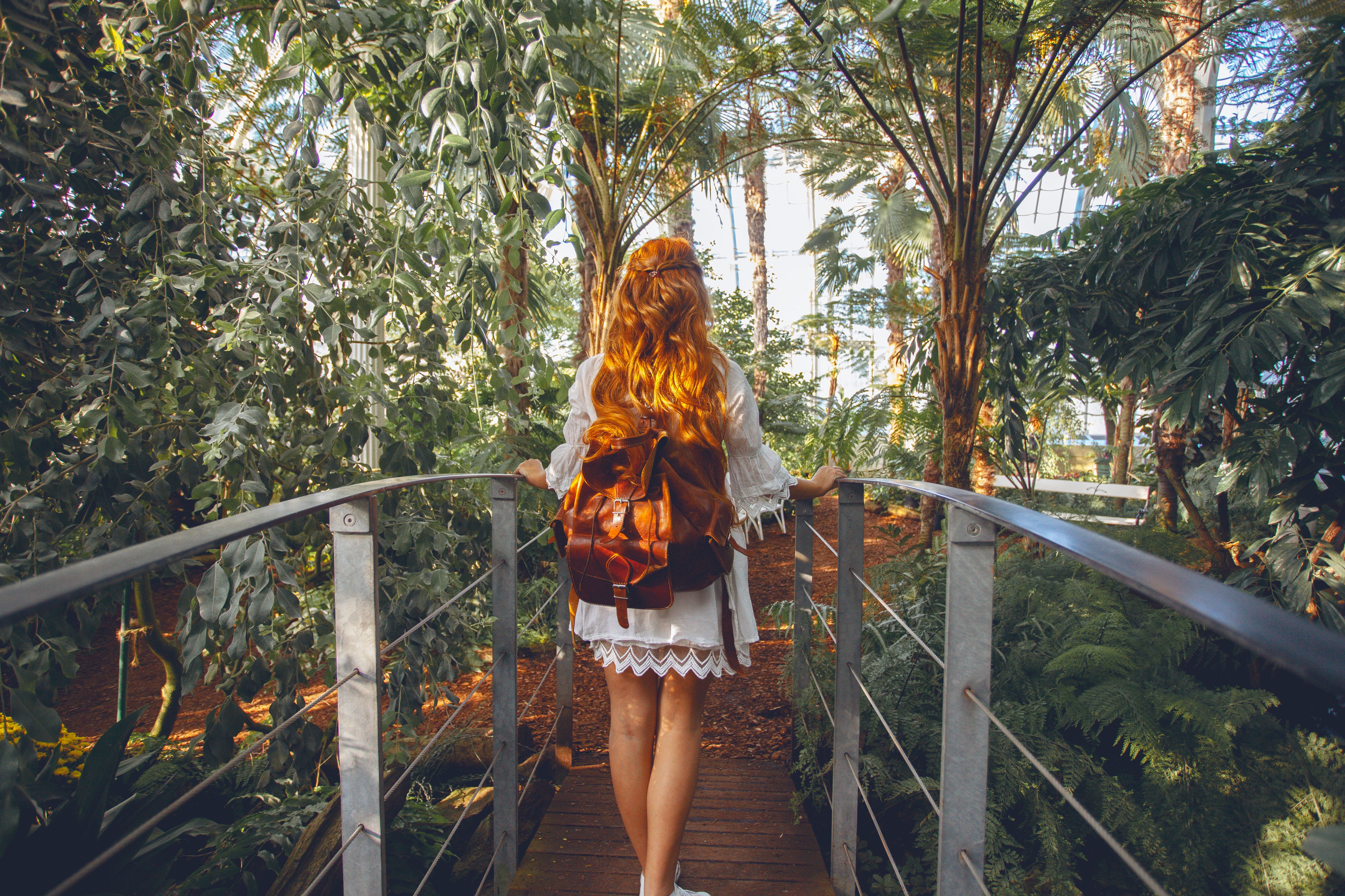 Frau mit Rucksack geht auf einer Holzbrücke durch einen üppigen, sonnendurchfluteten tropischen Botanischen Garten.