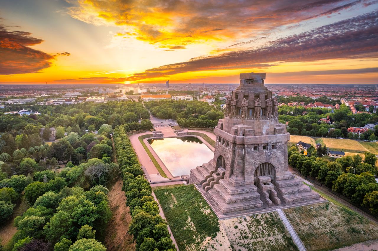 Luftaufnahme des Völkerschlachtdenkmals in Leipzig bei Sonnenuntergang, umgeben von grünen Parks und reflektierendem Wasserbecken, mit farbenfrohem Himmel und weitem Stadtblick im Hintergrund.