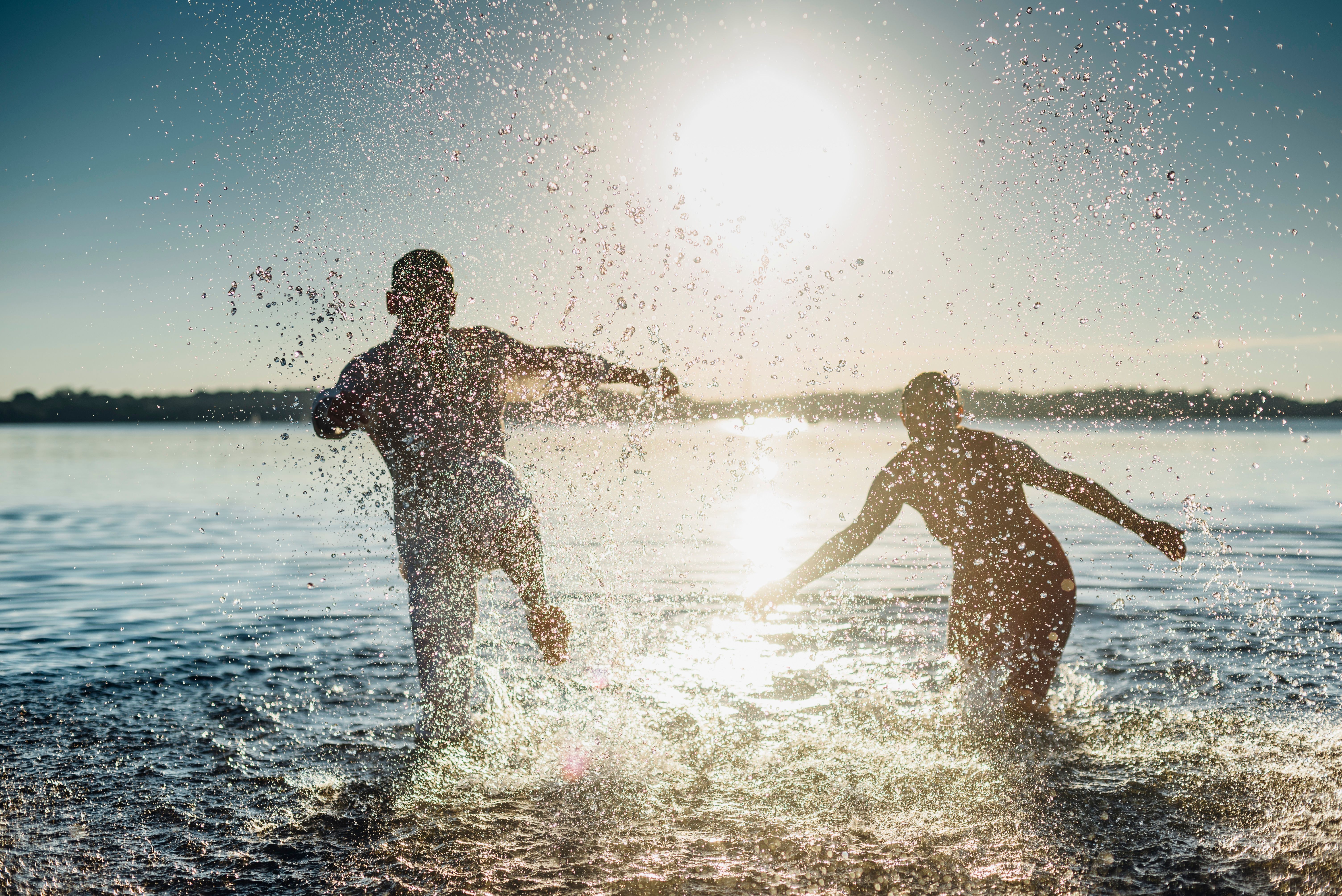 Zwei Personen springen lachend ins Wasser bei Sonnenuntergang, umgeben von spritzenden Wasserfontänen, die eine lebendige und freudige Atmosphäre am Seeufer vermitteln.