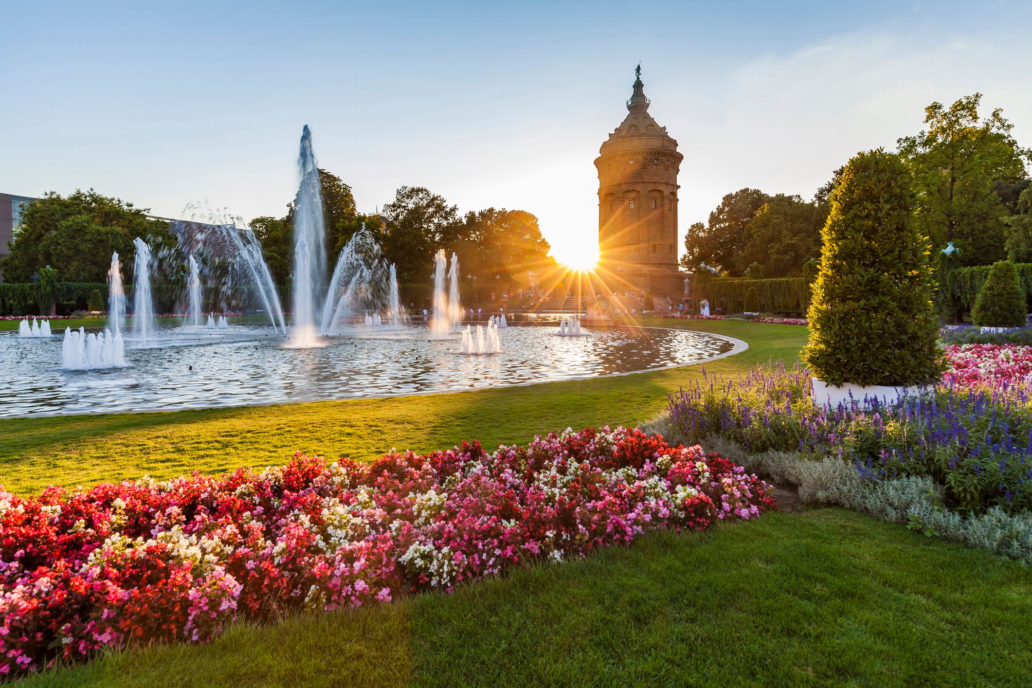 Sonnenuntergang hinter historischem Wasserturm, umgeben von einem großen Springbrunnen und farbenfrohen Blumenbeeten in gepflegtem Park.