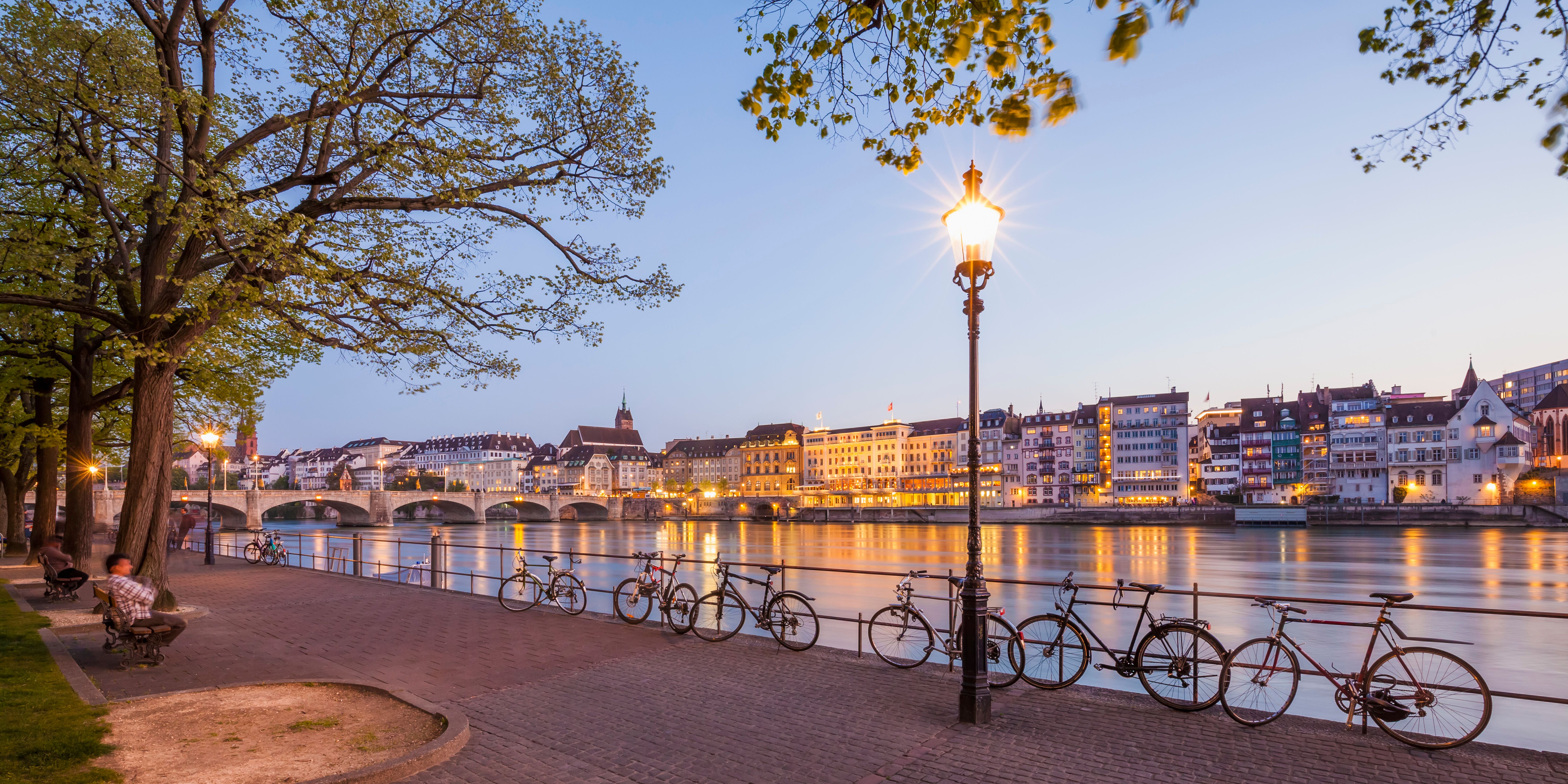Uferpromenade am Fluss mit Fahrradständer und daran geparkten Fahrrädern, beleuchtete Stadt mit historischen Gebäuden und Brücke im Hintergrund bei Dämmerung.