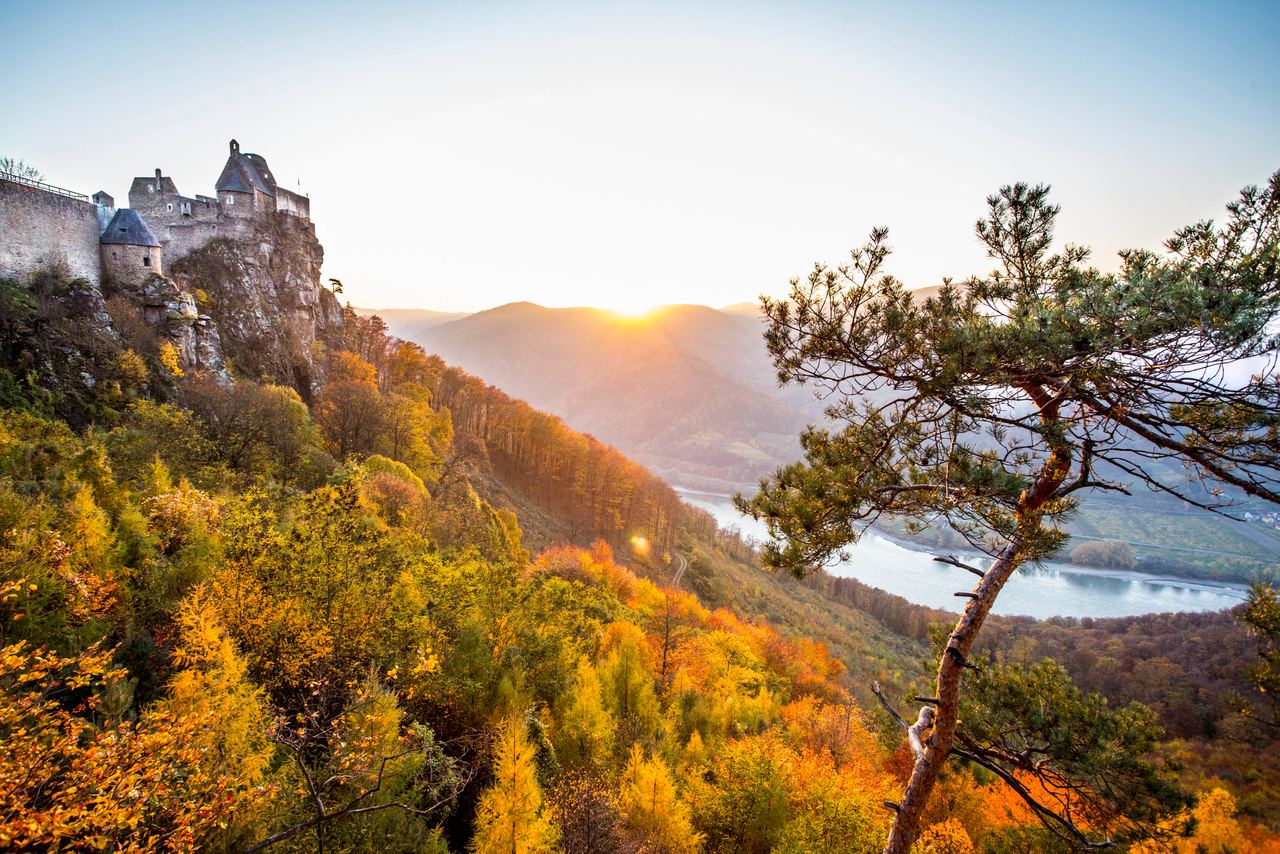 Malerische Aussicht auf eine Burg auf einem bewaldeten Hügel bei Sonnenuntergang, mit bunten Herbstbäumen und einem Fluss im Tal.