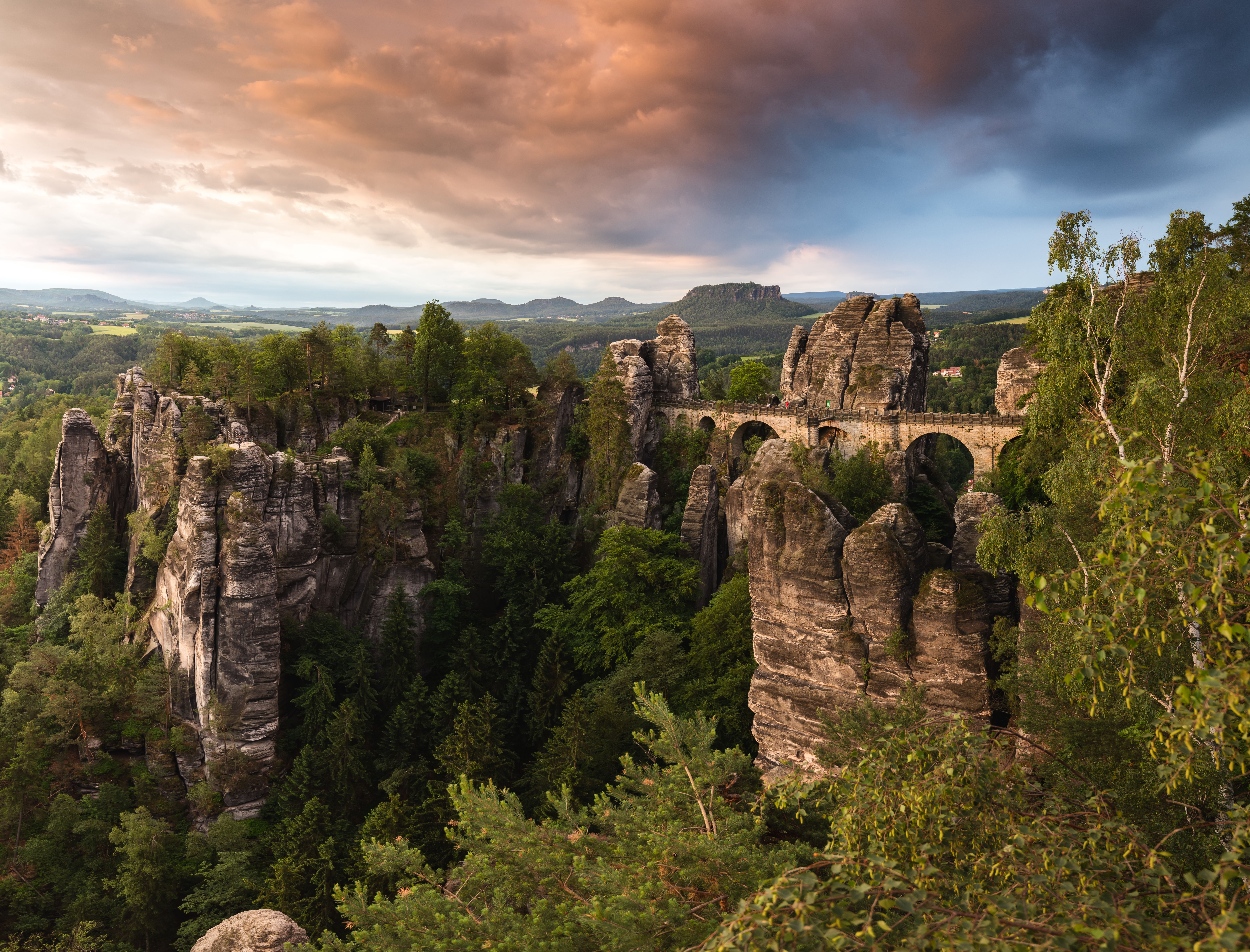 Felsformationen der Sächsischen Schweiz mit der Basteibrücke, umgeben von dichtem Wald, unter dramatischem Abendhimmel. Panoramablick auf die einzigartige Landschaft und natürliche Schönheit der Region.
